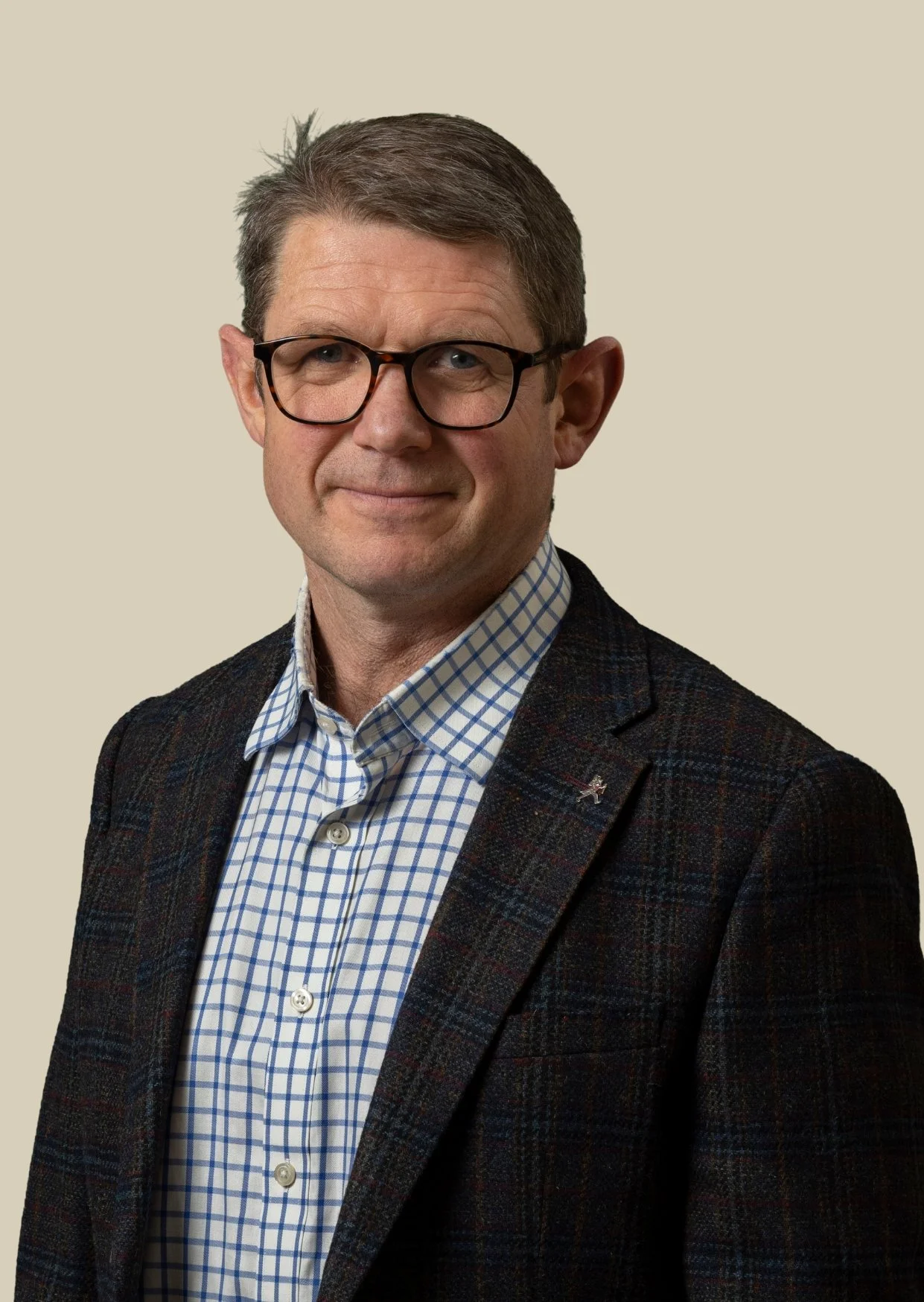 A man with short brown hair, glasses, wearing a checked shirt and a dark blazer with a small pin, standing against a light beige background.