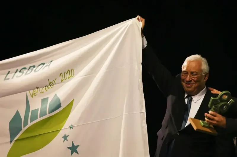 A man in a suit holding a plaque and smiling while holding a flag that reads "Velledor 2020," with a logo featuring a stylized green leaf and blue stars.