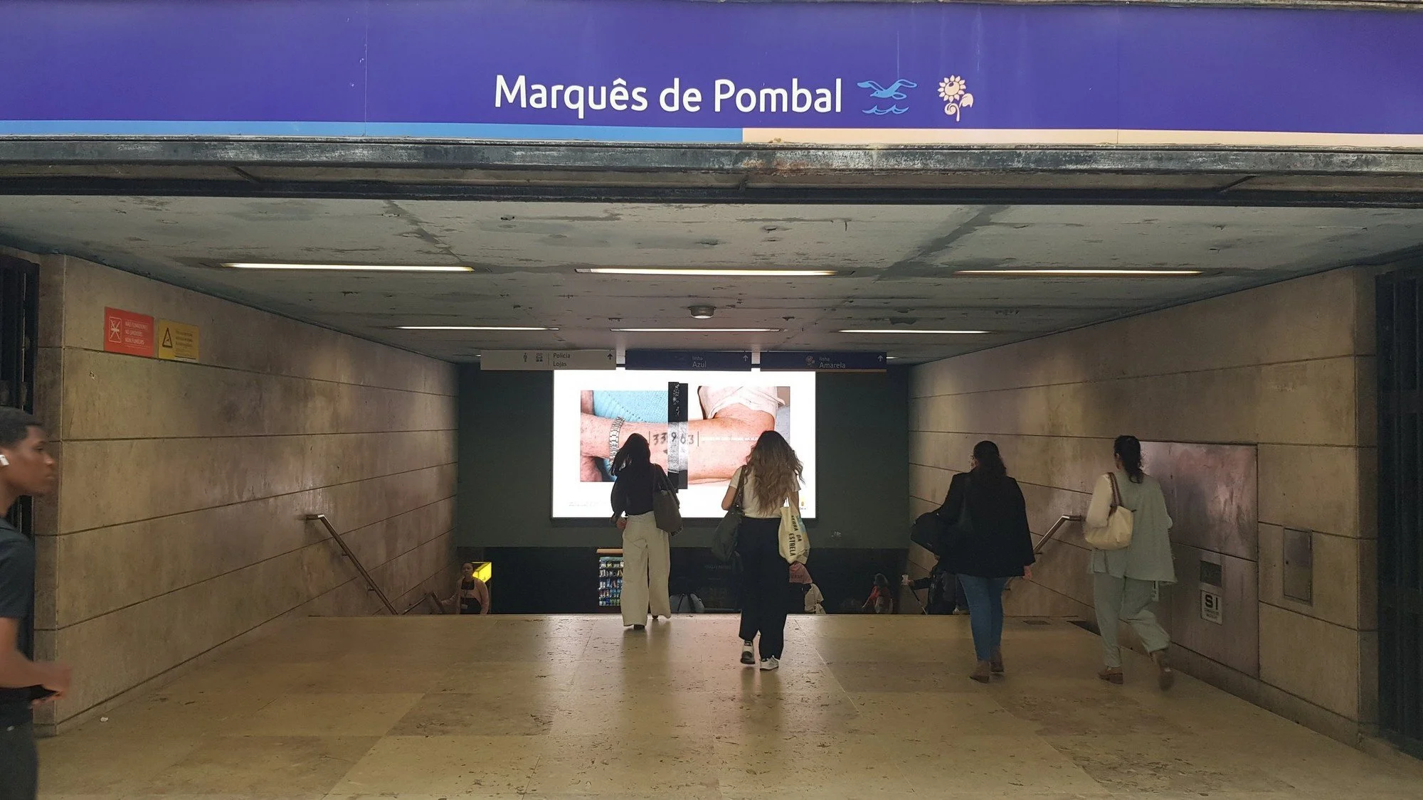 People walking in the underground Marquês de Pombal metro station with a large digital advertisement screen on the wall showing a close-up of arms and a measuring tape.