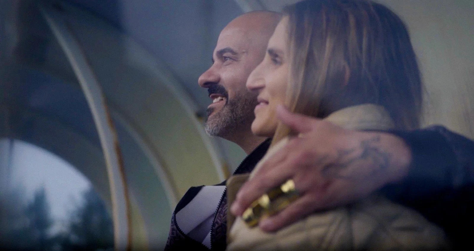 Two people, a man with a beard and a woman with long hair, are sitting close together, smiling and enjoying each other's company.