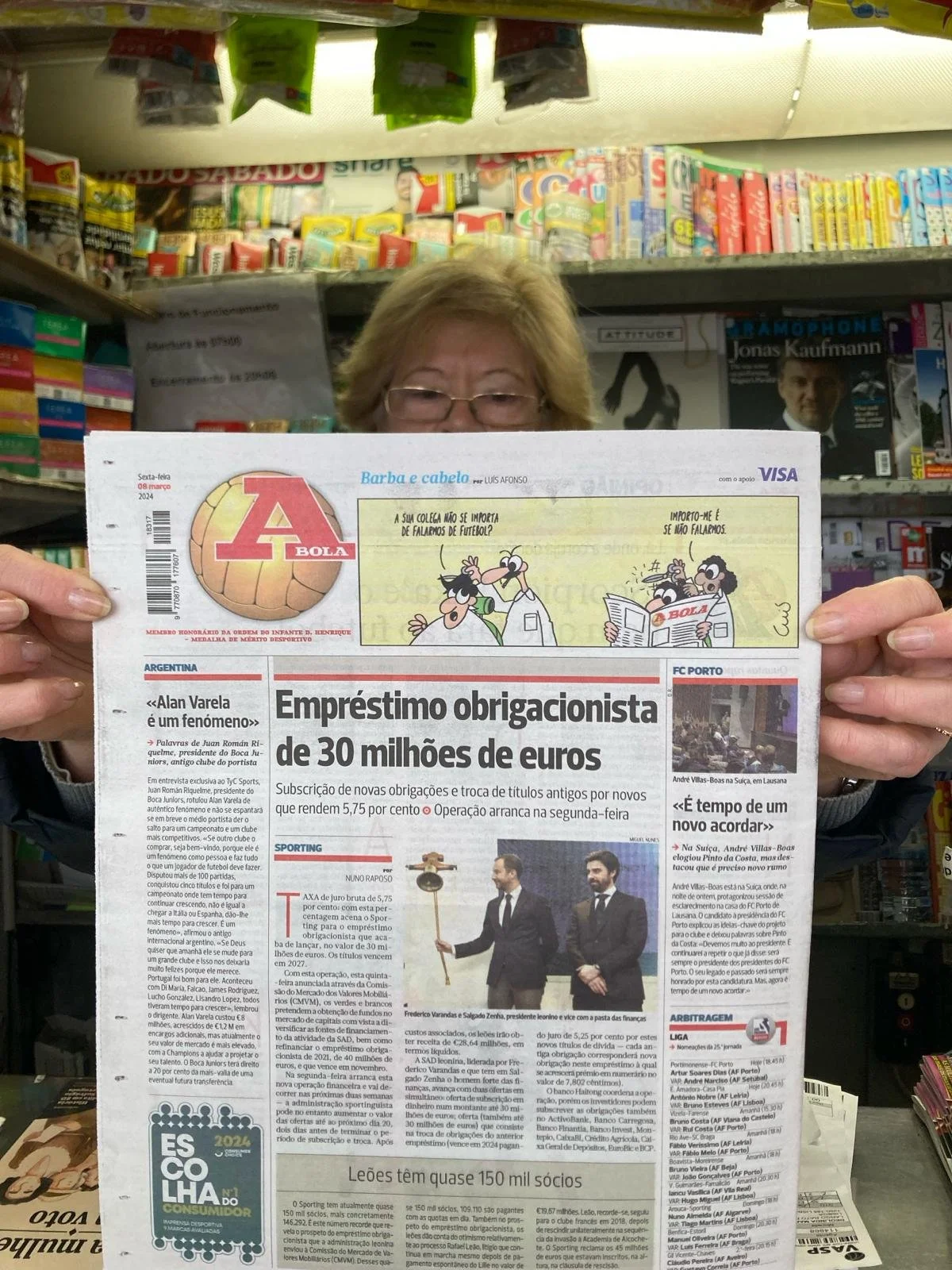Woman holding a newspaper in a store aisle filled with snacks and magazines.