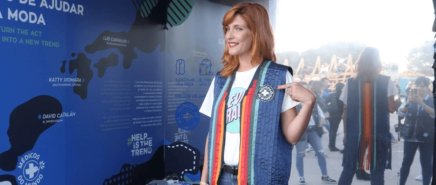 Woman with red hair wearing a t-shirt and rainbow-striped vest pointing to her badge at a health aid event.