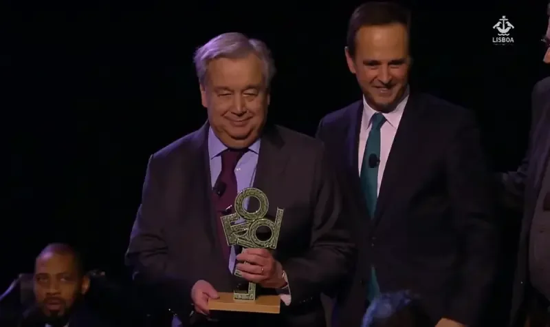 Two men in suits on stage, one holding an award, smiling, with a dark background and a logo in the top right corner.