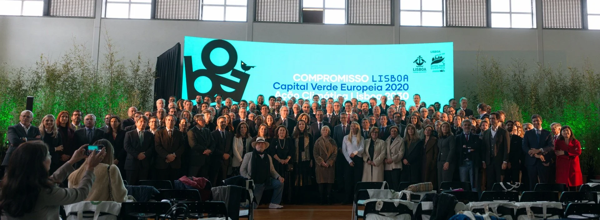 Group of people attending a conference or event in front of a large screen displaying the text "Compromisso Lisboa Capital Verde Europeia 2020 Ação Climática Lisboa 2030" with logos of Lisbon and Portugal, in an indoor venue with banners and green plants.
