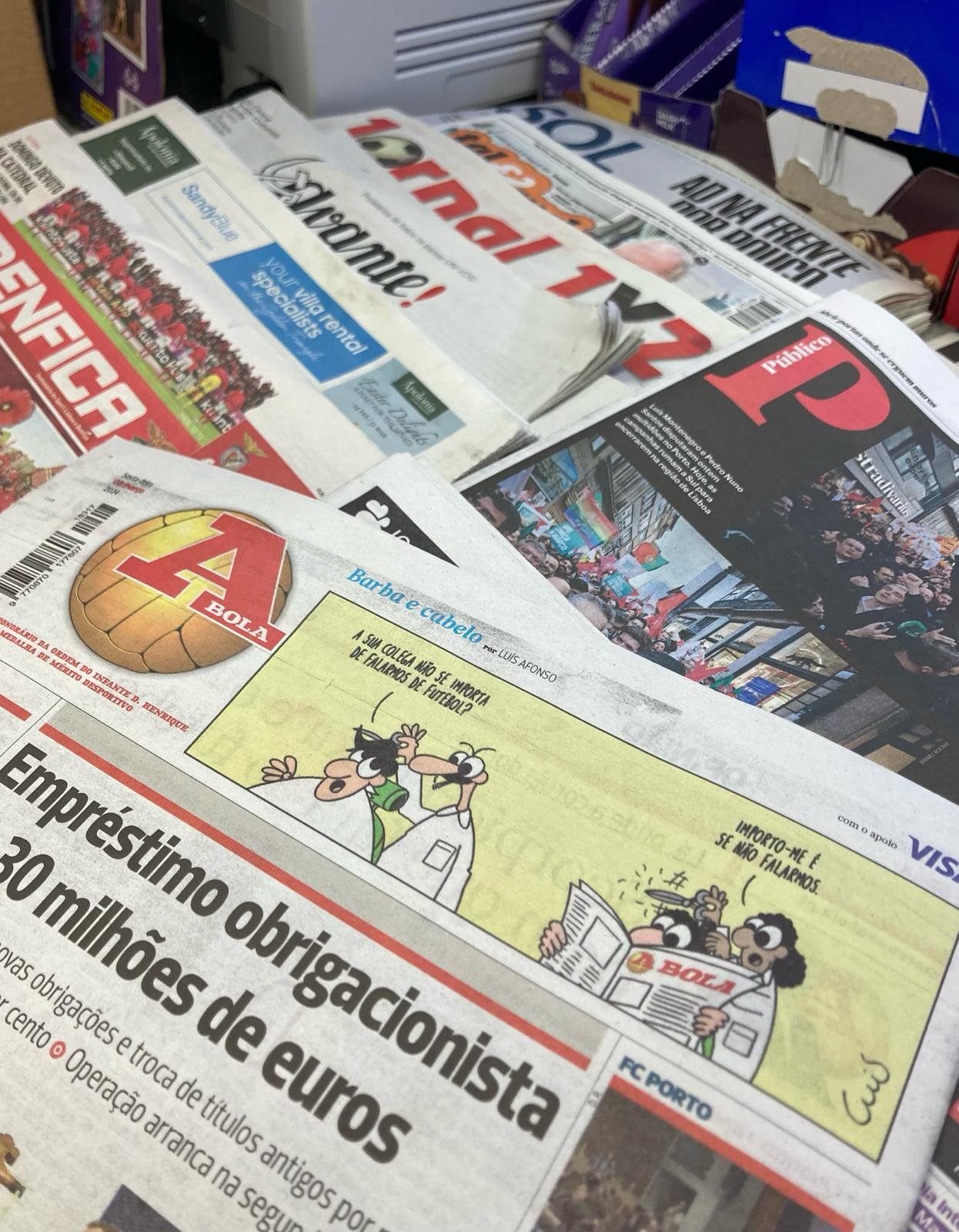 A collection of Portuguese newspapers and magazines displayed on a table, including a comic strip featuring cartoon characters and headlines about soccer and current events.