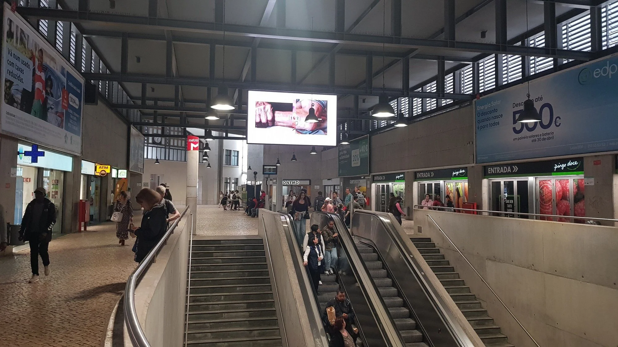Interior of a busy train or metro station with people moving around, some on escalators, some walking, and advertisements and signs visible on the walls.