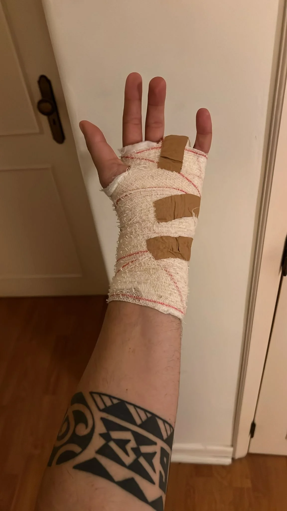 A person's arm with a tattoo, wrapped with medical bandages and brown adhesive tape, raising their hand indoors against a plain wall and a door.