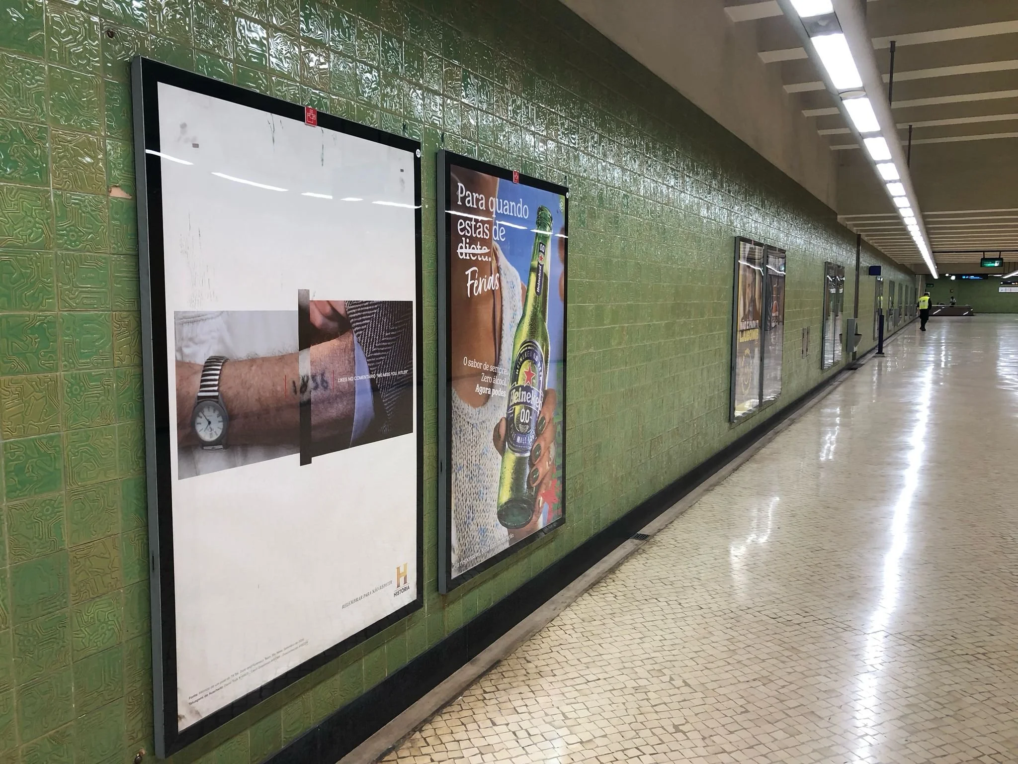 Underground subway corridor with a green tiled wall decorated with framed advertisement posters, and a person walking in the distance.