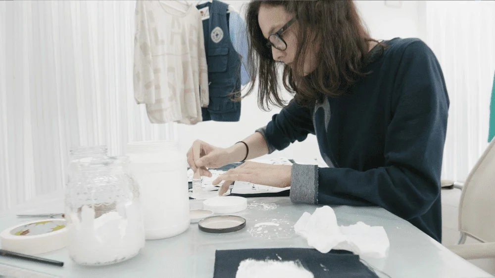 A woman with glasses and long hair, wearing a dark jacket, working with white powder on a table, surrounded by jars, containers, and cloths in a bright room.