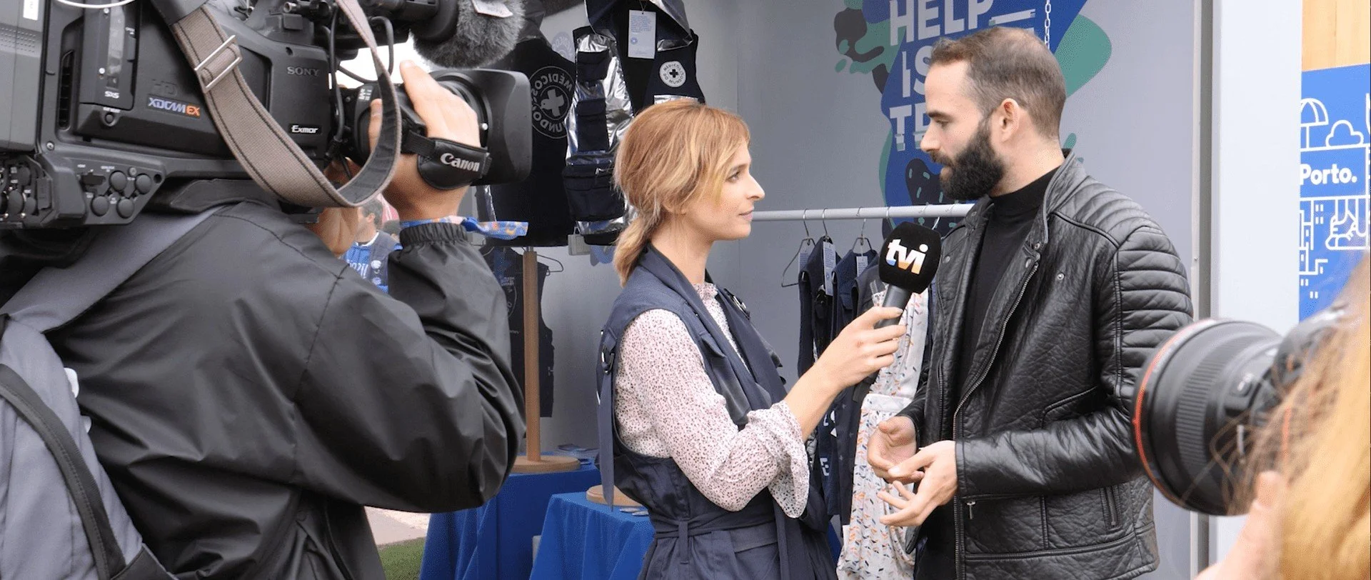 A woman interviews a man with a TVI microphone at an outdoor event, with a person filming and making a video recording nearby.