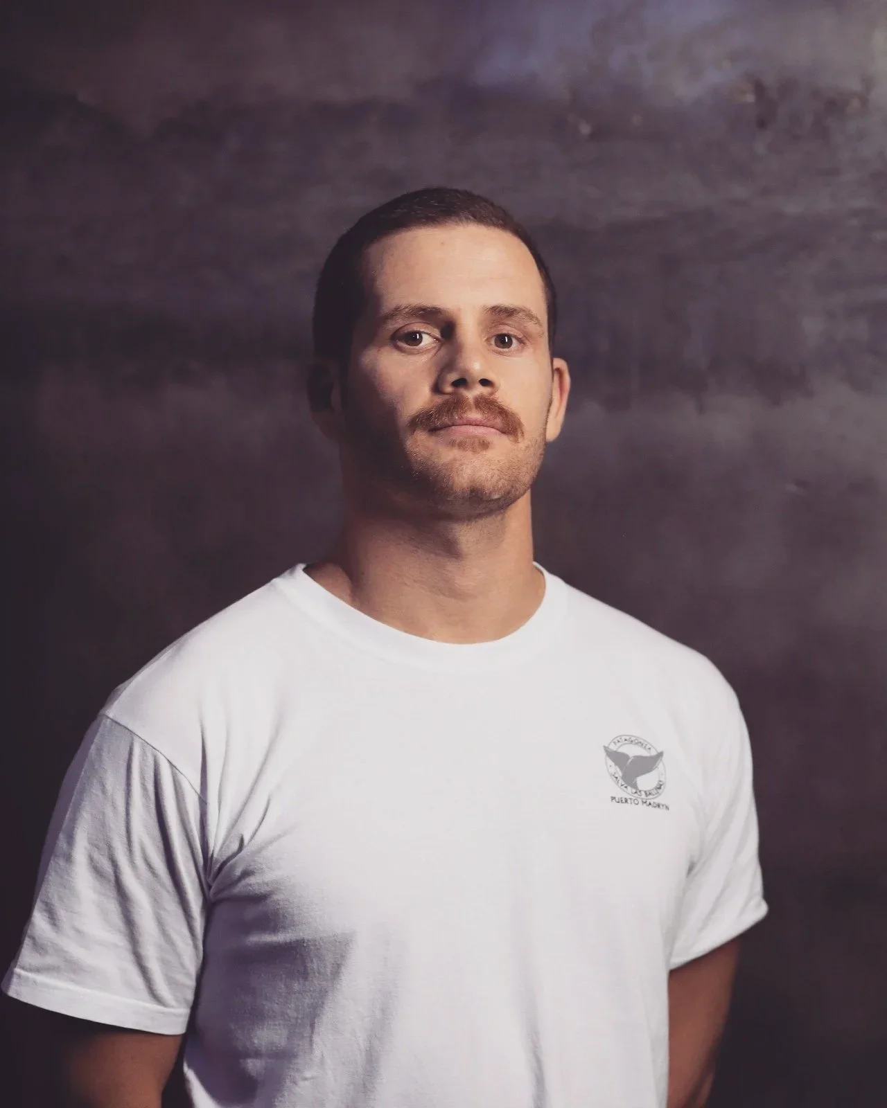 A man with short dark hair and a mustache wearing a white t-shirt standing against a dark, textured background.