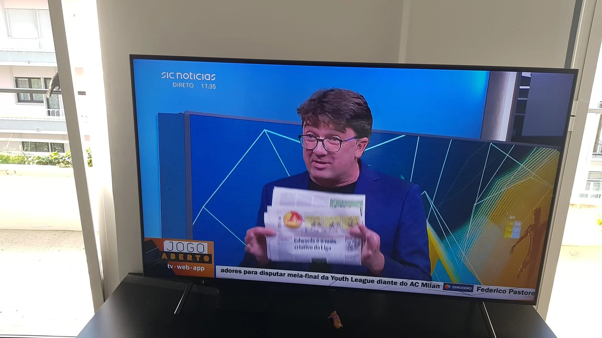 A man with glasses and dark hair on a TV screen holding a newspaper, with news graphics and text in Portuguese, and a balcony with a building visible in the background.