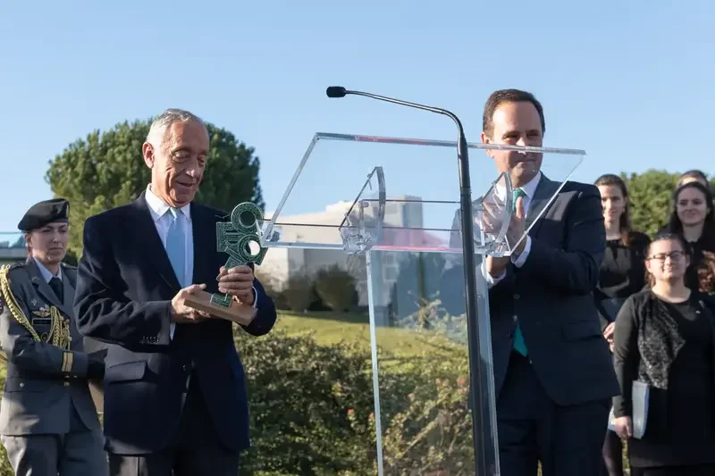 Two men in suits at a podium, one holding a trophy and the other clapping, during an outdoor awards ceremony with several women standing in the background.