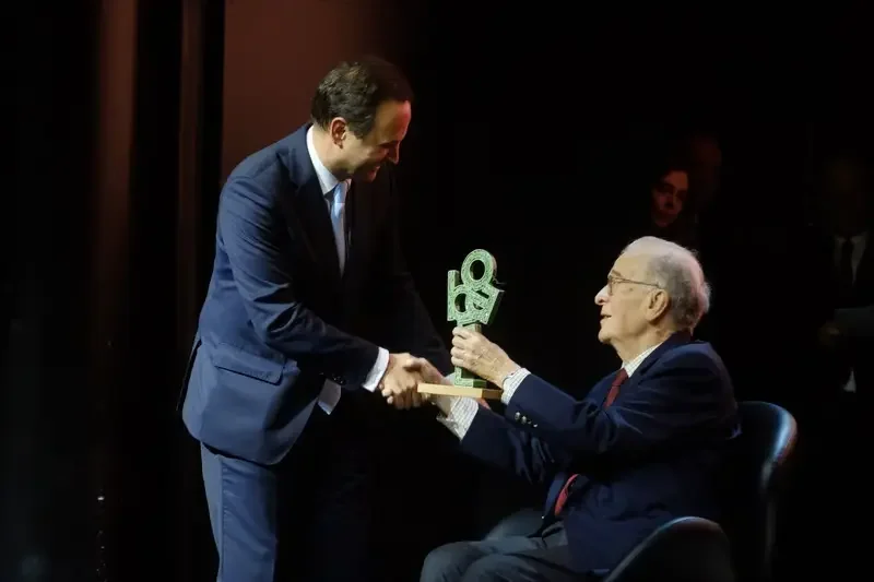 A man in a suit shaking hands with an elderly man in a suit, who is sitting and receiving an award on a stage.