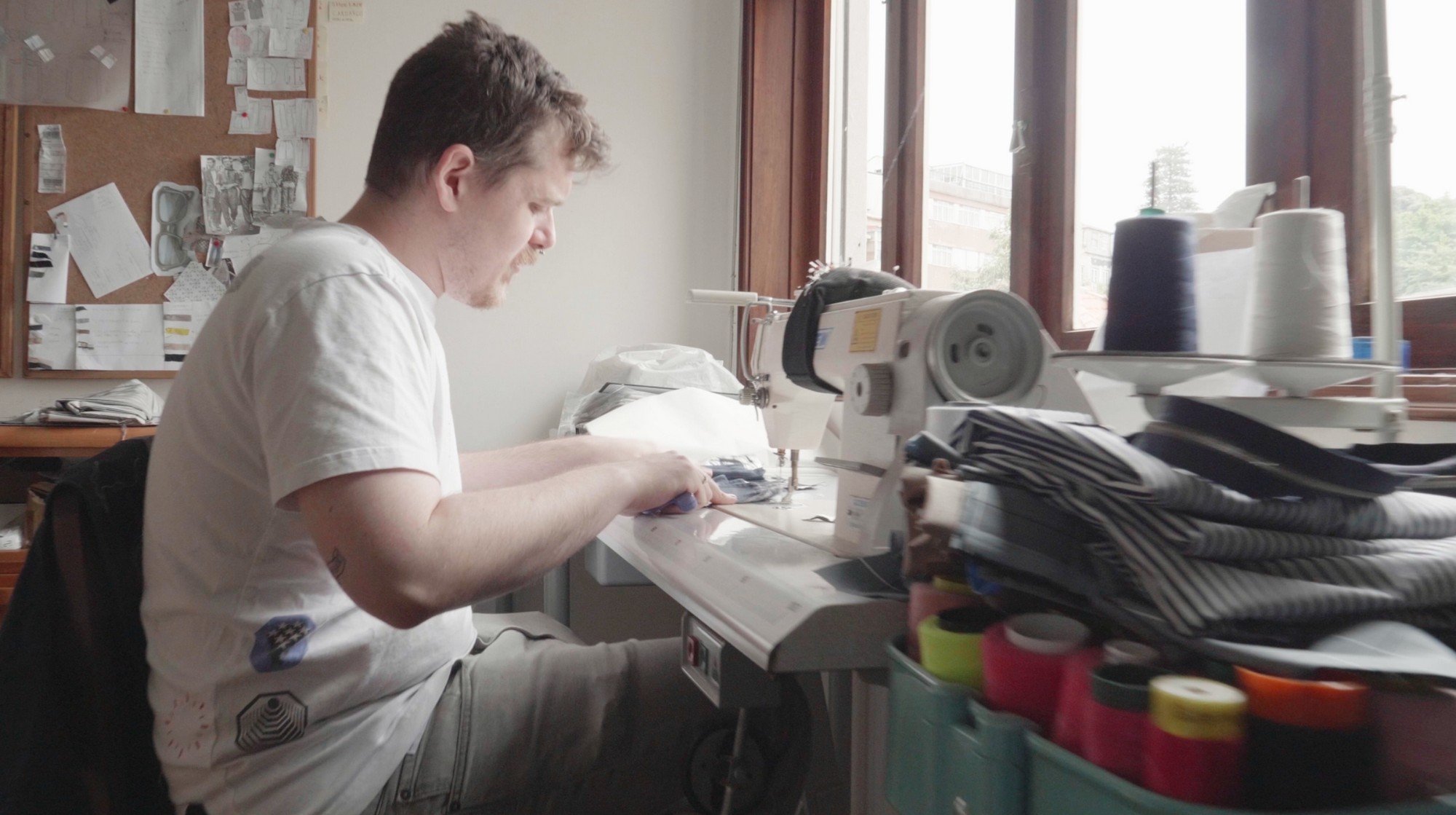 Young man working at a sewing machine in a well-lit room with tools and fabric around.