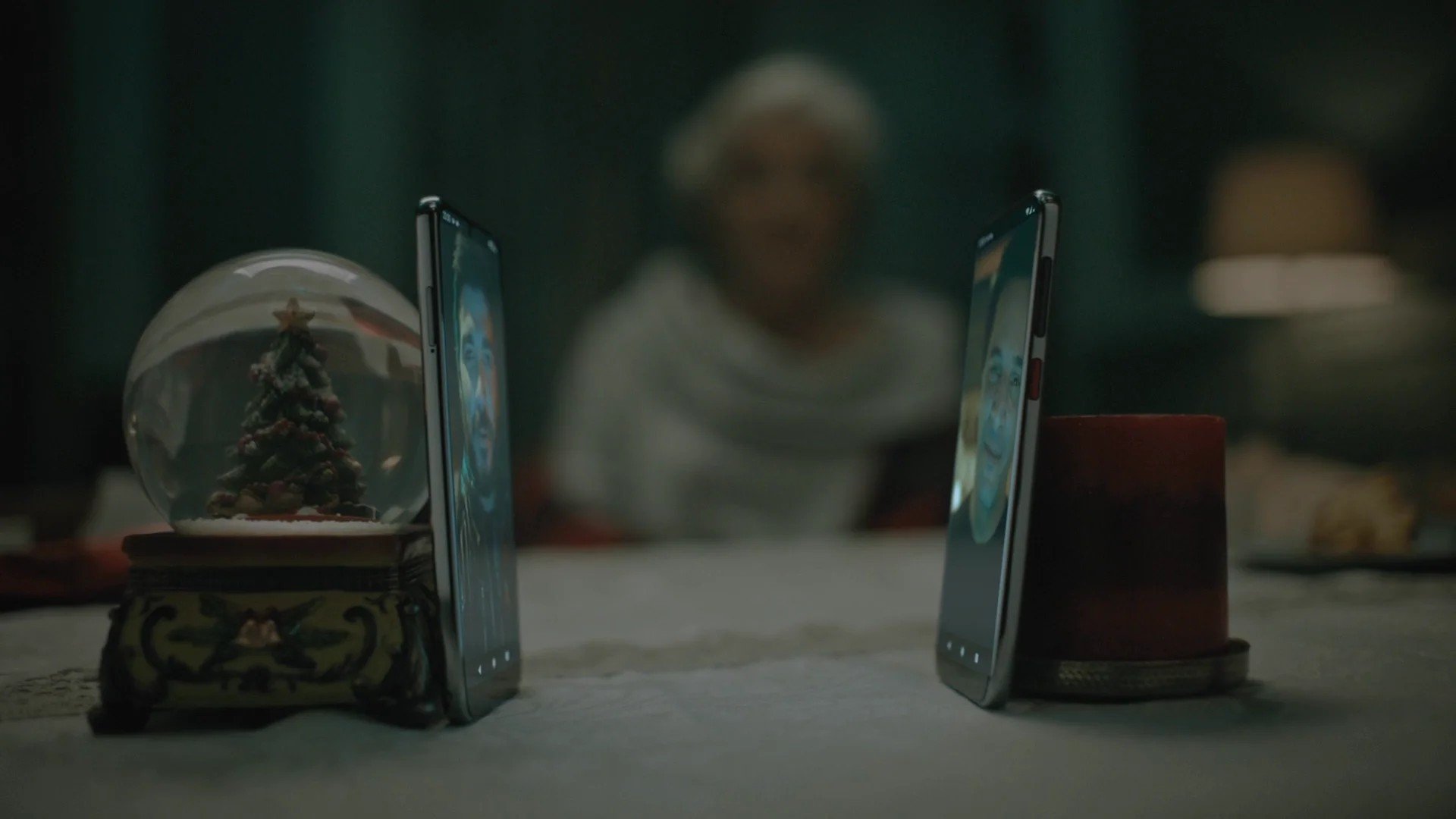 Two smartphones on a table facing each other, with a snow globe featuring a decorated Christmas tree on a stand on the left and a red cup on the right. In the background, a person with short gray hair is blurred and smiling.