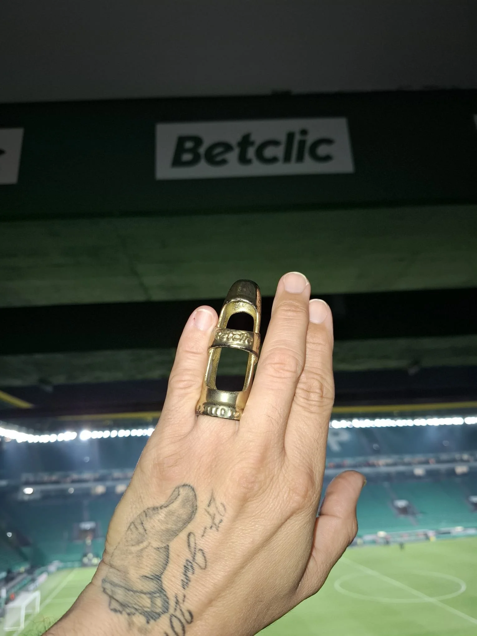 A person's hand with a tattoo and a large gold ring showing, touching a green sign with the word 'Betclic' in a stadium.