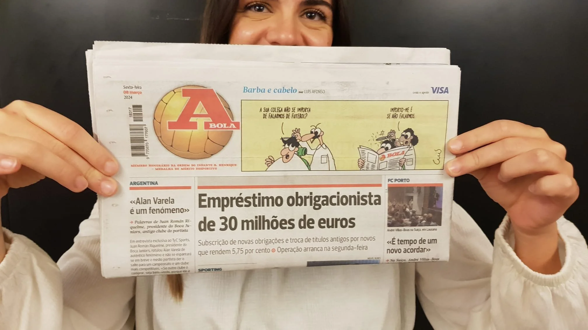 Person holding a newspaper titled 'A Bola' with a comic strip at the top and a headline about a 30 million euro sports loan, wearing a white sweater, against a black background.