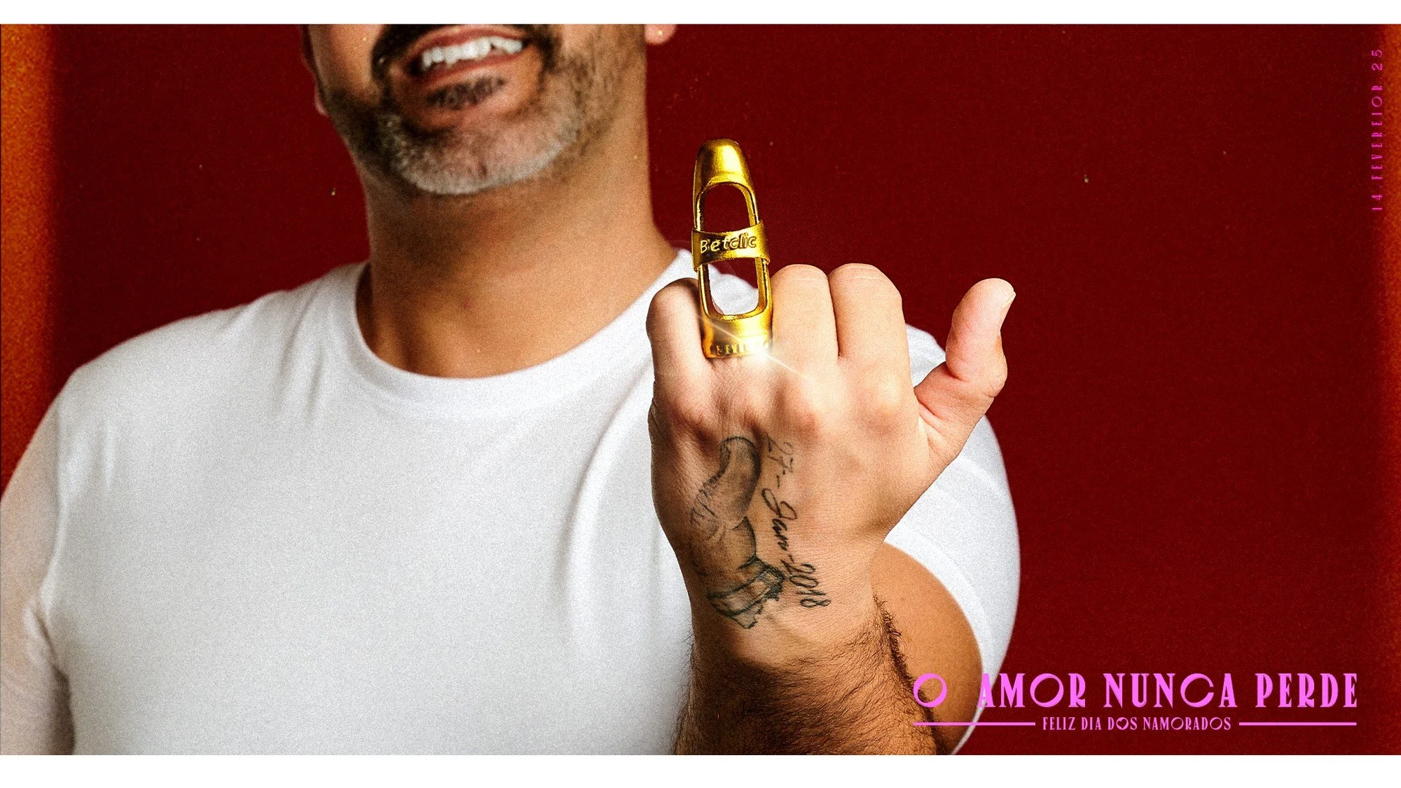 A man with a beard and tattoos on his hand, wearing a white t-shirt, stands in front of a red background. He is holding up his hand with a gold ring shaped like a carabiner that has the word 'Béatific' on it. The image includes pink text in the bottom right corner that reads 'O AMOR NUNCA PERDE FELIZ DIA DOS NAMORADOS'.