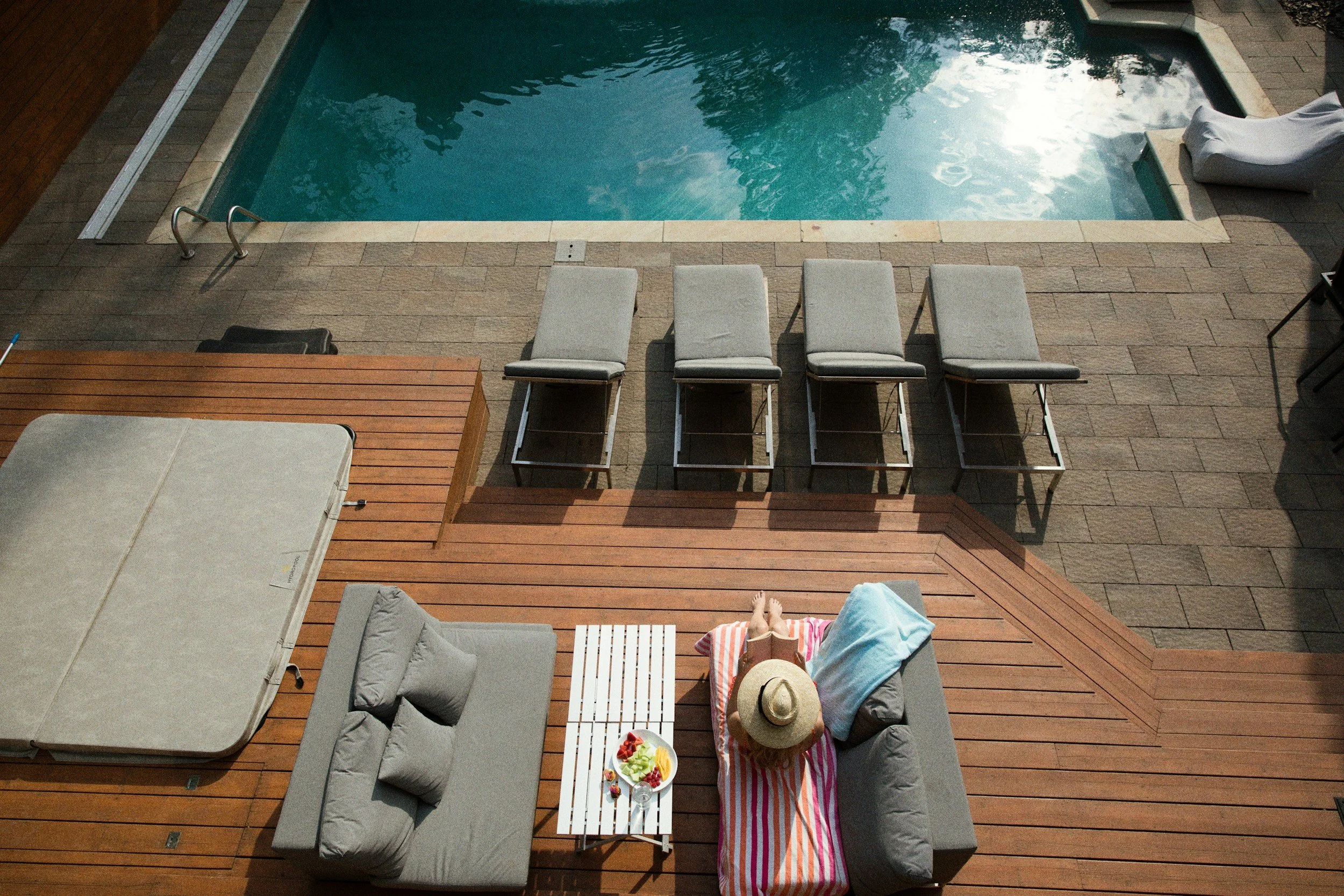 A person lying on a lounge chair next to a pool, wearing a hat and colorful towel, with a plate of food nearby, surrounded by other lounge chairs and outdoor furniture on a wooden deck.