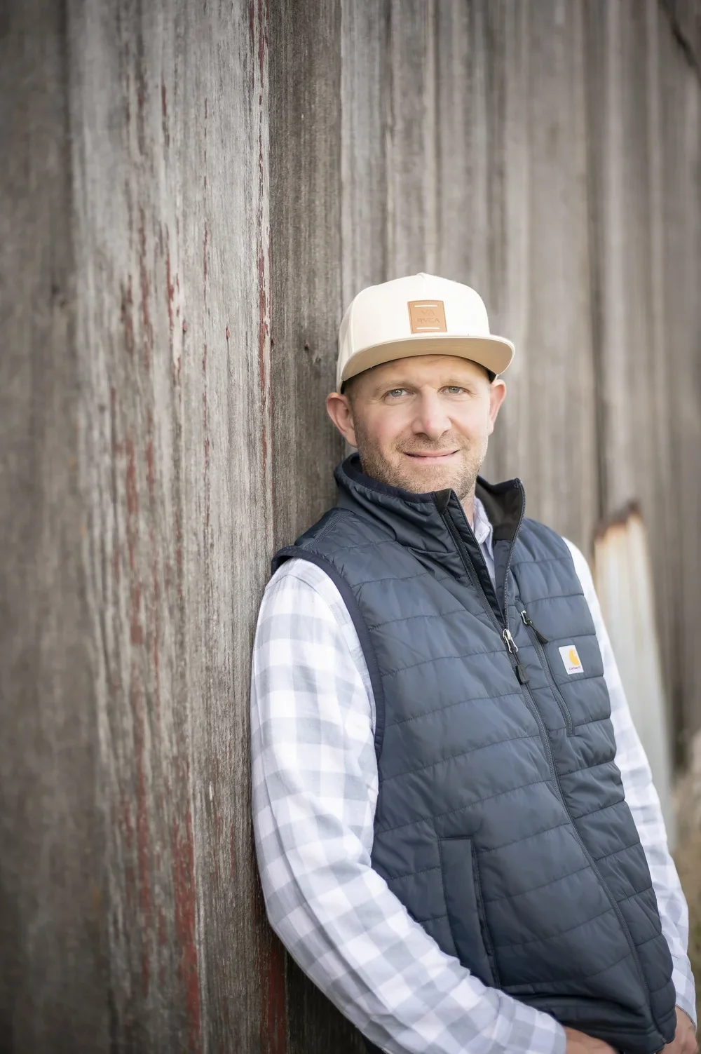 A man leaning against a weathered wooden wall, wearing a beige cap, a navy blue Carhartt vest, and a checkered shirt.