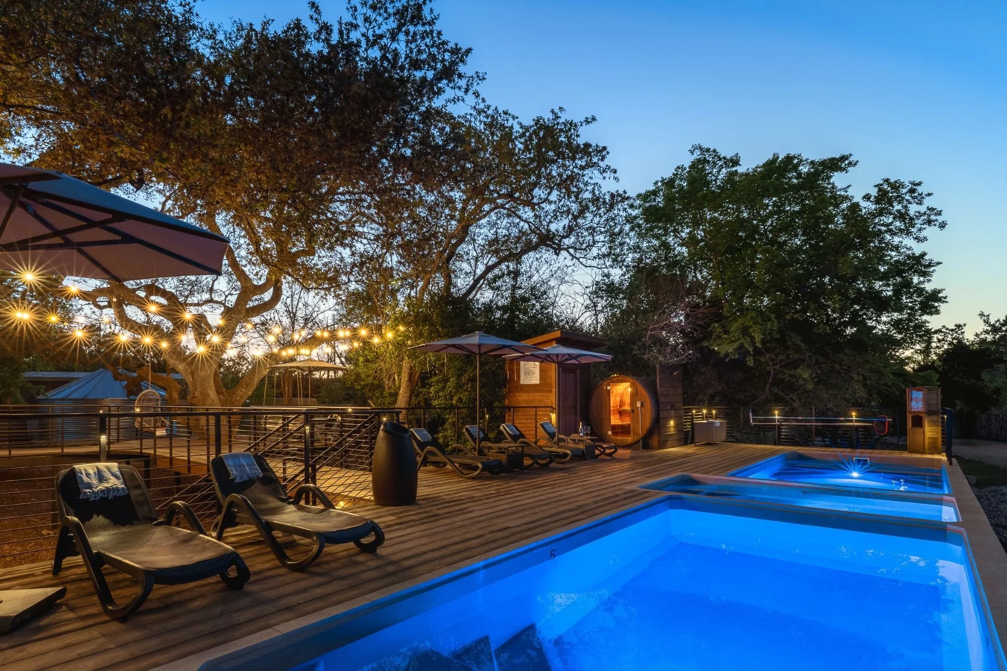 St. Louis. Missouri, Tide and Timber Luxury Outdoor Living Spaces specializing in decks, covered patios, pools. Outdoor swimming pool area at dusk with lounge chairs, umbrellas, string lights, trees, and a sauna in the background.