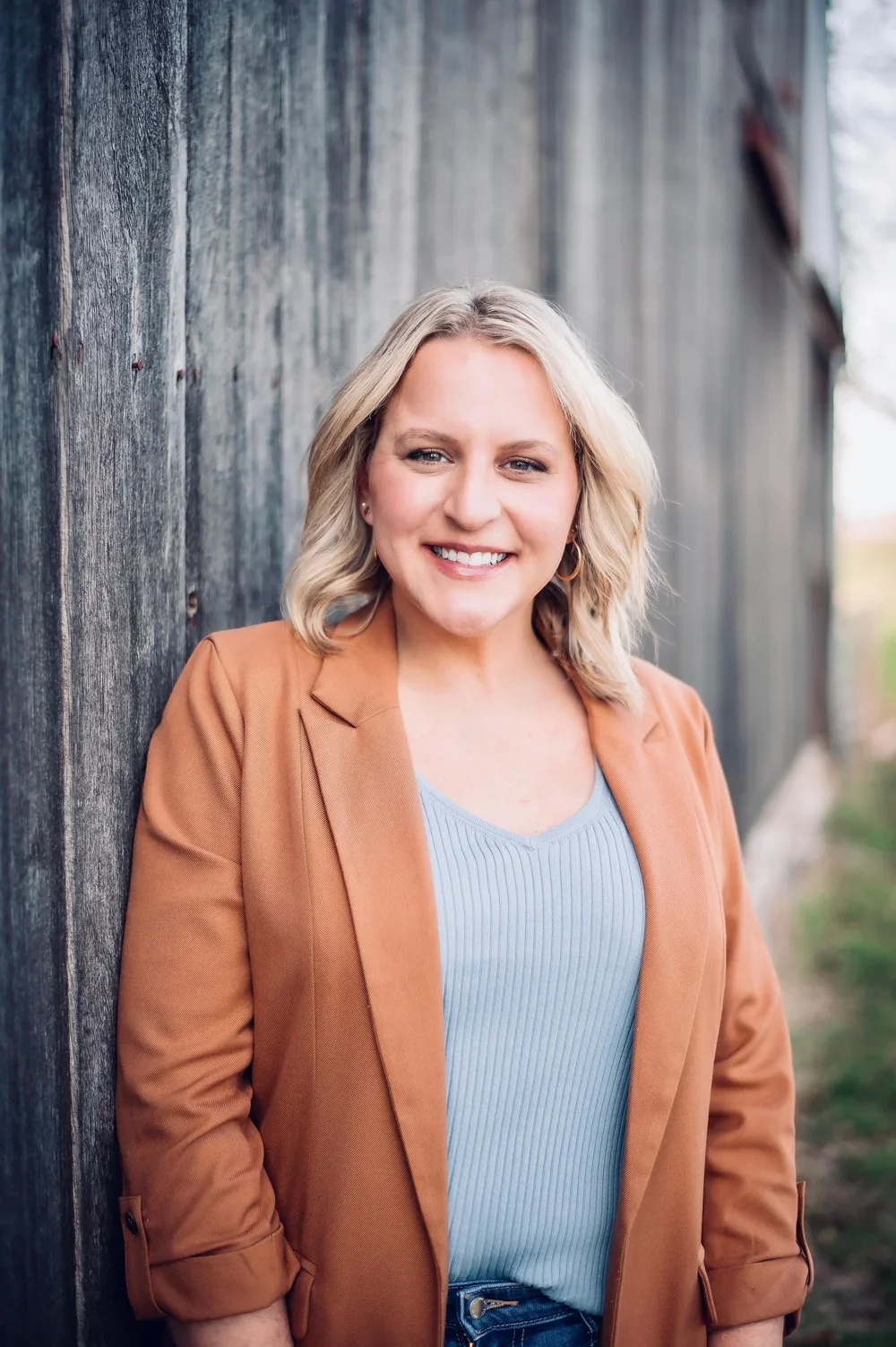 A smiling woman with shoulder-length blonde hair wearing a tan blazer over a light blue ribbed top, standing against a weathered wooden wall outdoors.