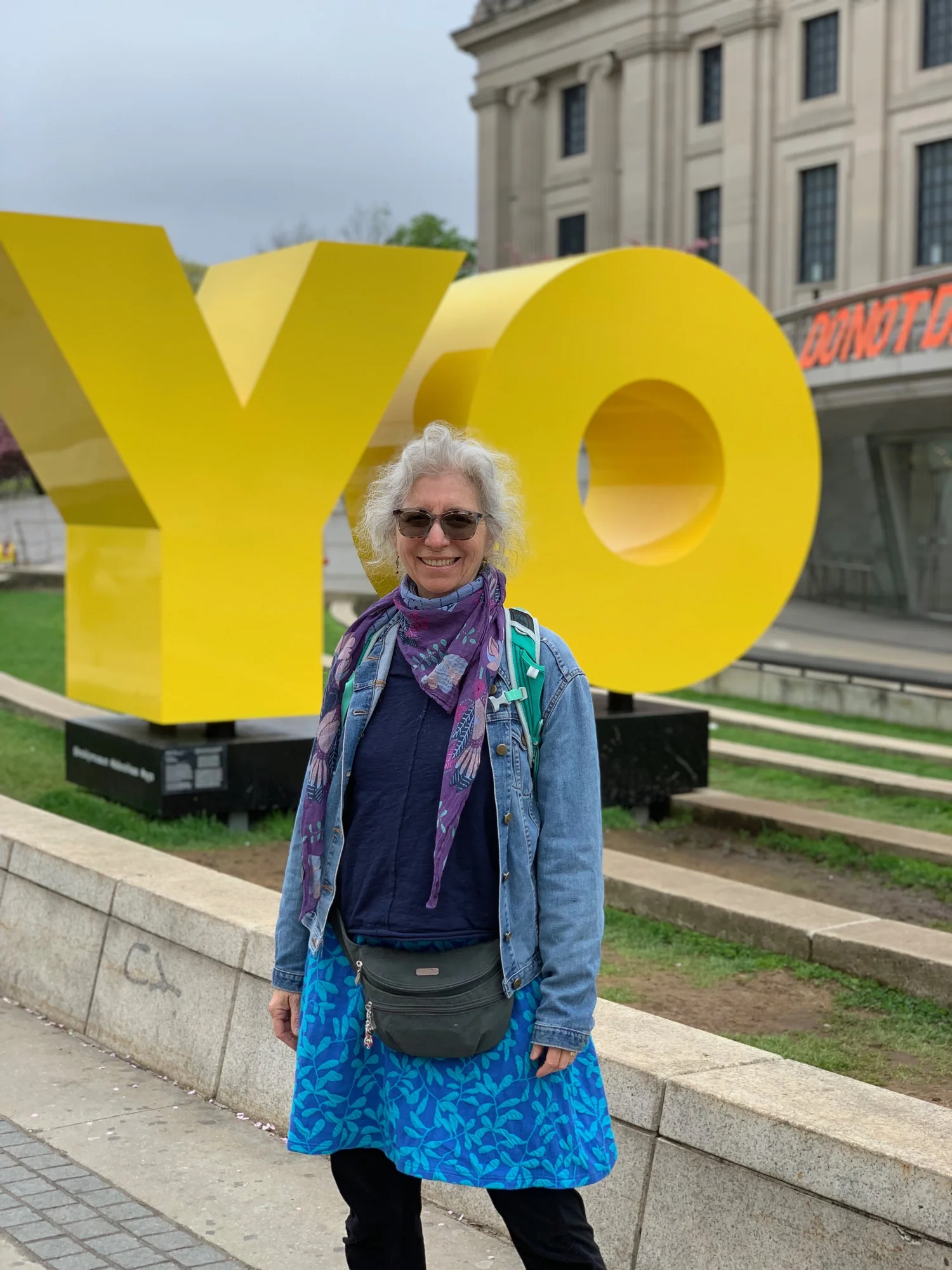 Smiling woman with curly gray hair, sunglasses, a denim jacket, a colorful scarf, and a blue skirt standing in front of a large yellow sculpture that spells "YQ" outdoors in an urban setting with a stone building in the background. Nancy DeNicolo