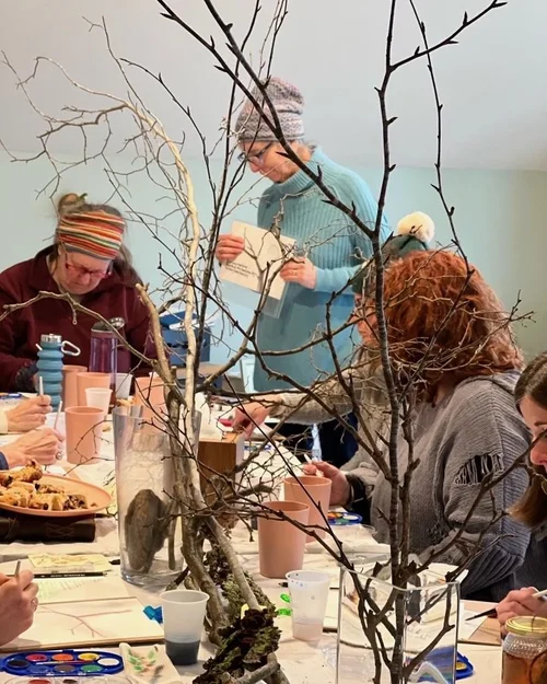 People sitting around a table creating art projects with branches and natural materials, indoors. Nancy DeNicolo Women's Art Circles and Experiences