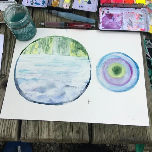 Watercolor paintings of a circular landscape scene and an abstract concentric circle pattern on white paper. Art supplies, including a jar of water, paintbrushes, and watercolor palettes, are visible on the table. Nancy DeNicolo Nature Watercolor
