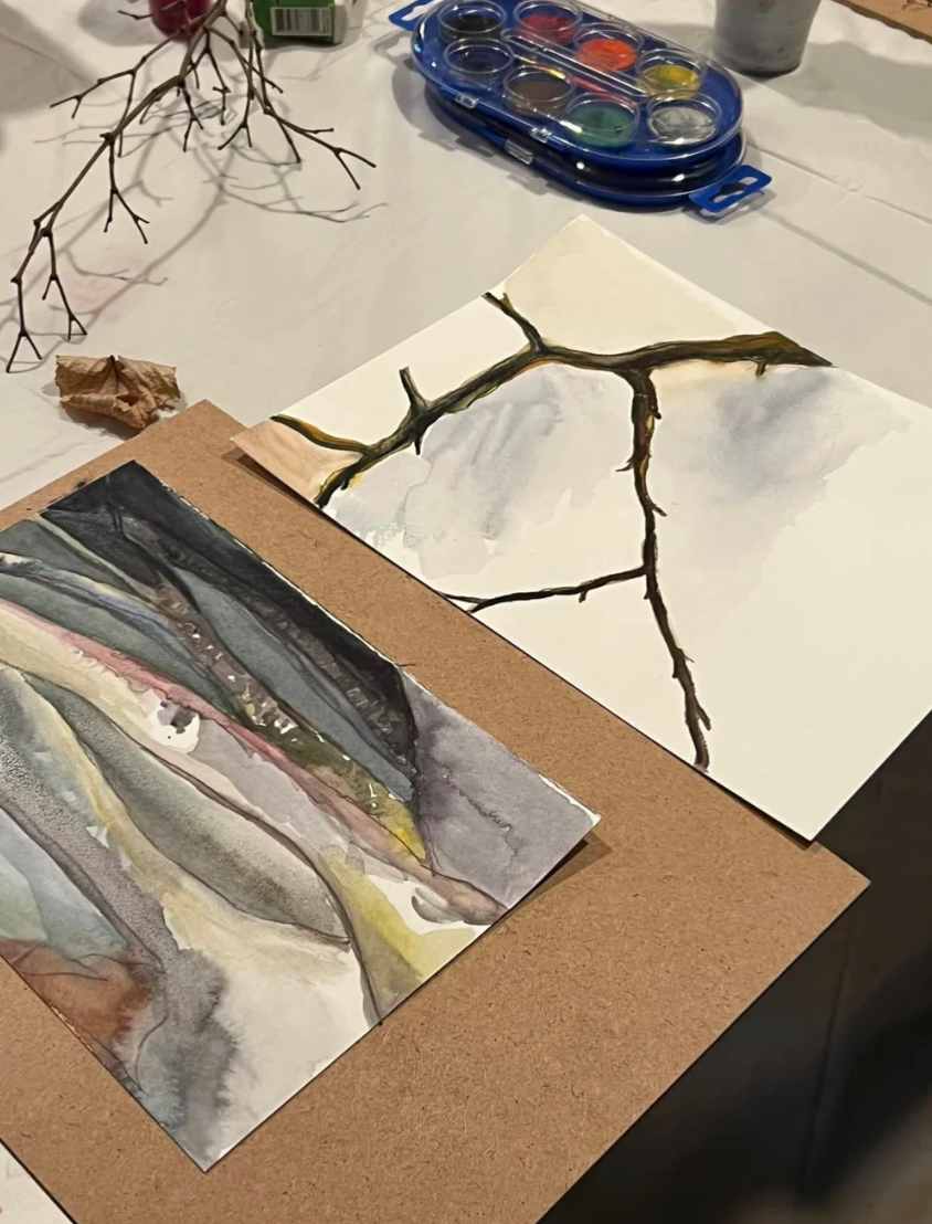 Two watercolor paintings of branches and landscapes are on a table surrounded by art supplies including a watercolor paint set, a dried branch, and a crumpled paper. Nancy DeNicolo Beginner Watercolor Classes Women's Circle