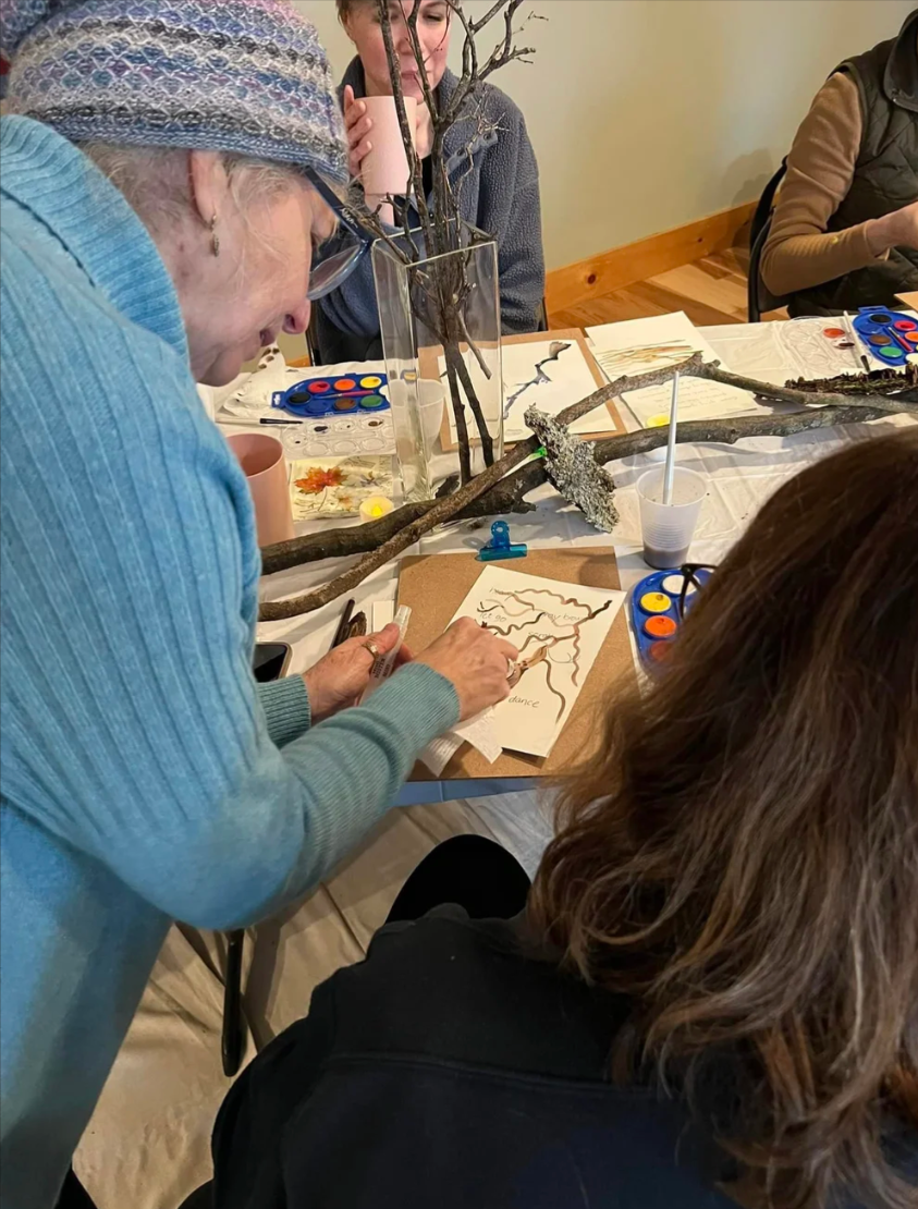 An elderly woman with glasses and a colorful knit hat working on an arts and crafts project at a table with several adults. The table has sticks, a vase with branches, paint, and paper with drawings. Nancy DeNicolo Watercolor classes for beginners