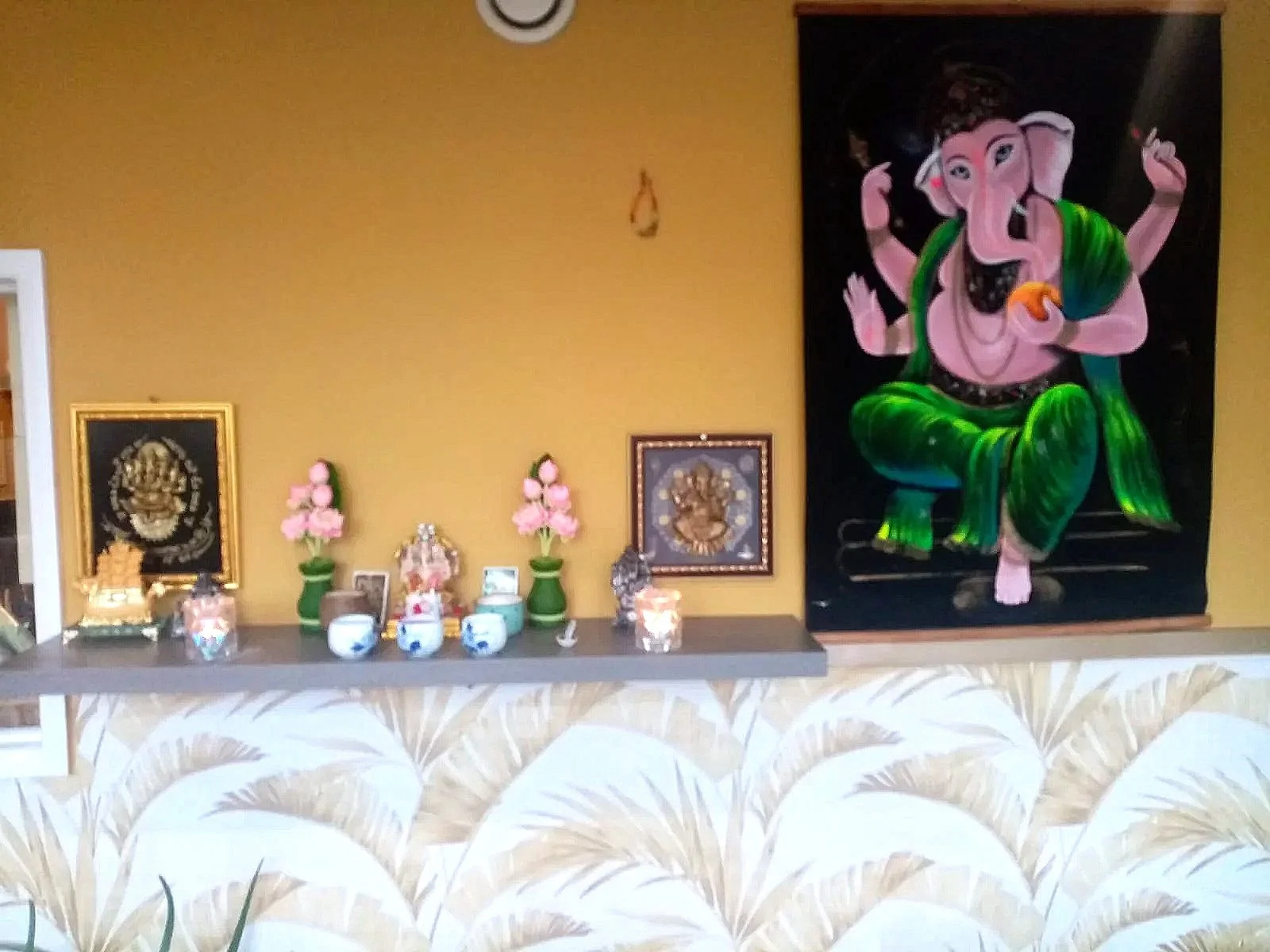 A decorated shrine with religious and cultural items, featuring a large colorful painting of Lord Ganesha on a black background, placed against a yellow wall.