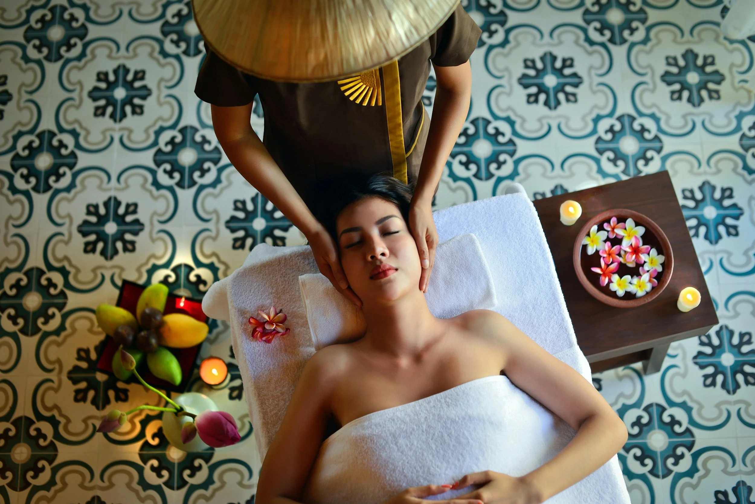 A woman receives a head massage while lying on a massage table in a spa room with floral and candle decorations.