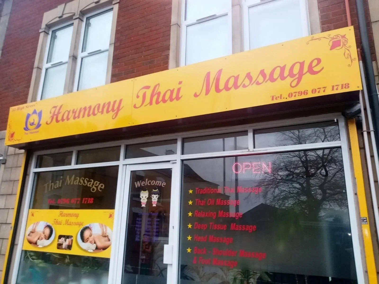 Yellow storefront sign for Harmony Thai Massage with pink lettering and phone number, window advertising massage services, neon 'OPEN' sign, promotional poster with images of massages, and list of massage types in red text.