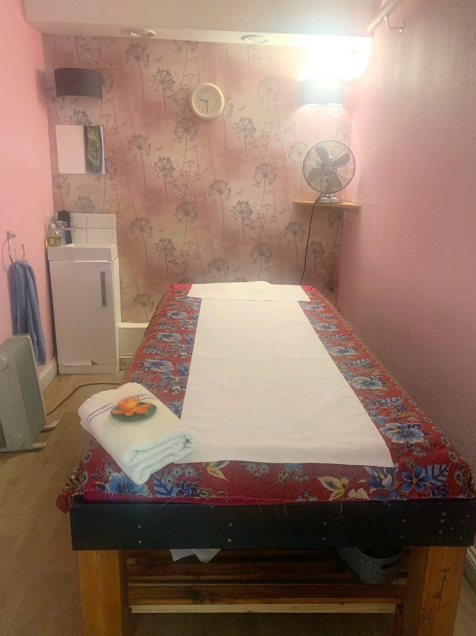 A massage table covered with white sheets and a towel, with a flower on top, in a room with pink floral wallpaper, a wall clock, a fan, and wall lights.