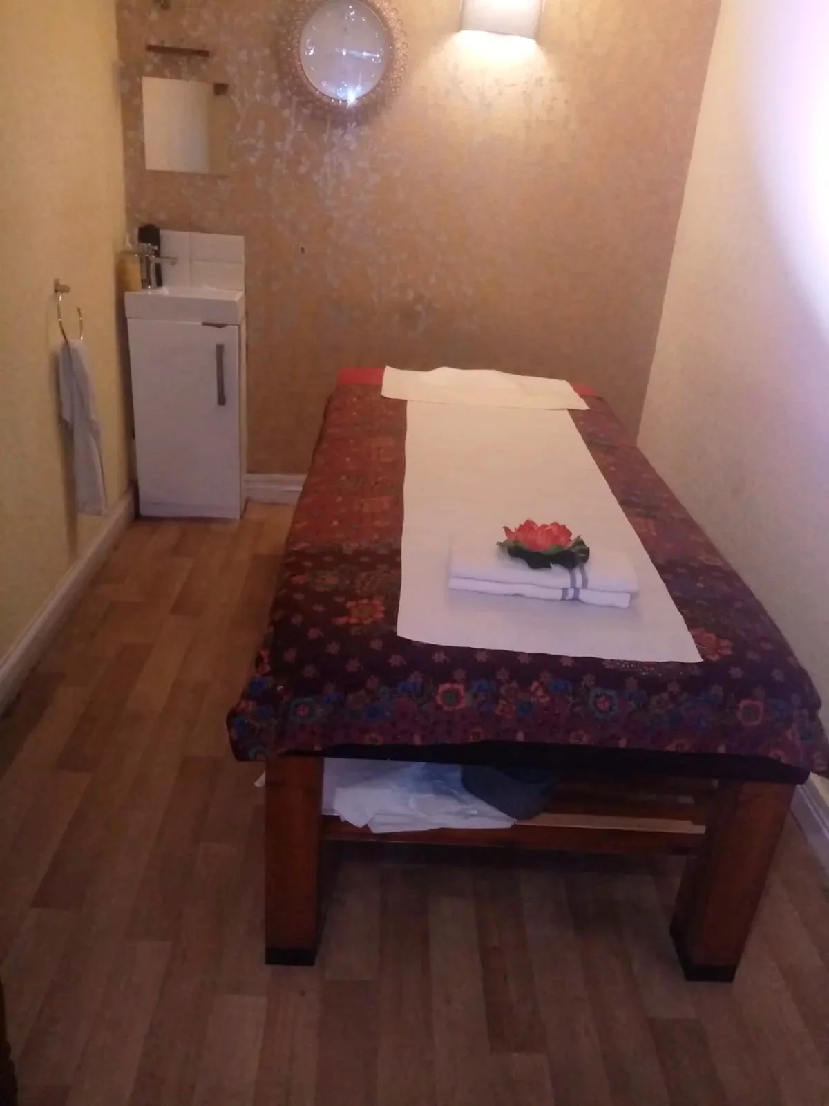 A small hotel room with a bed, a towel folded with a flower on top, a mini sink, a mirror, a wall-mounted light, and a mirror reflecting the ceiling. The bed has a floral bedspread and is arranged with a white sheet and pillow.