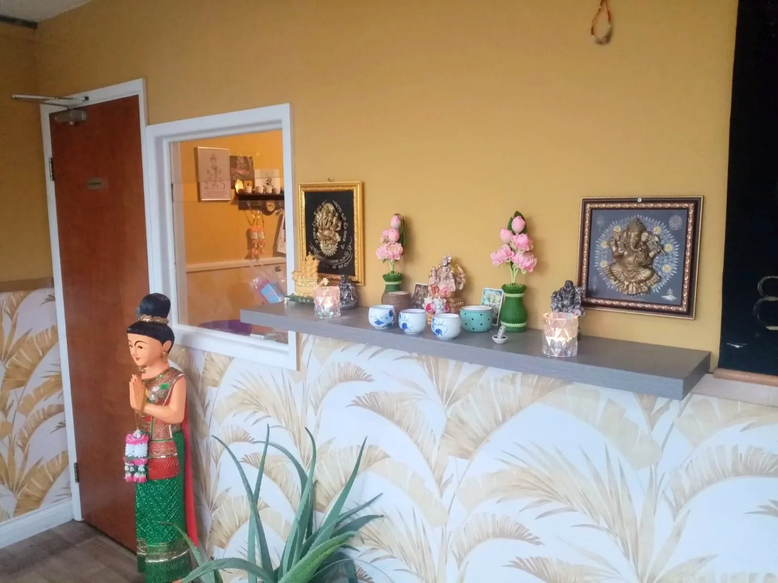 Decorative Hindu altar with framed images, flower vases, and statues, including a standing deity figurine, against a yellow wall with pineapple-patterned wallpaper.