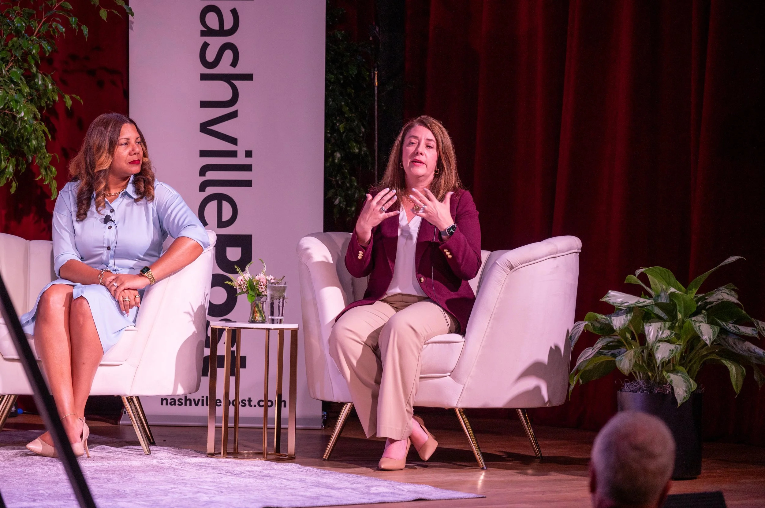 Two women sit on white armchairs on a stage, engaged in a discussion.