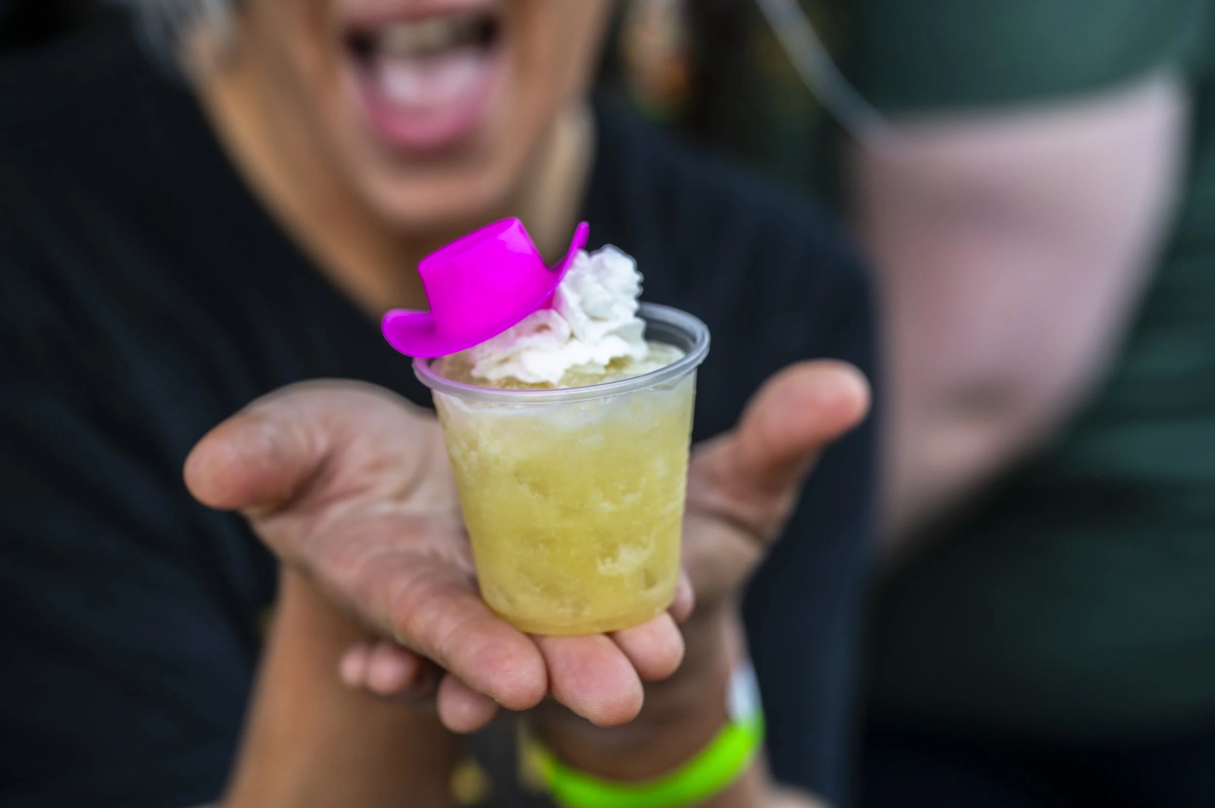 Person holding a yellow frozen margarita topped with whipped cream and a small pink hat decoration in a clear plastic cup, smiling with mouth open in the background.