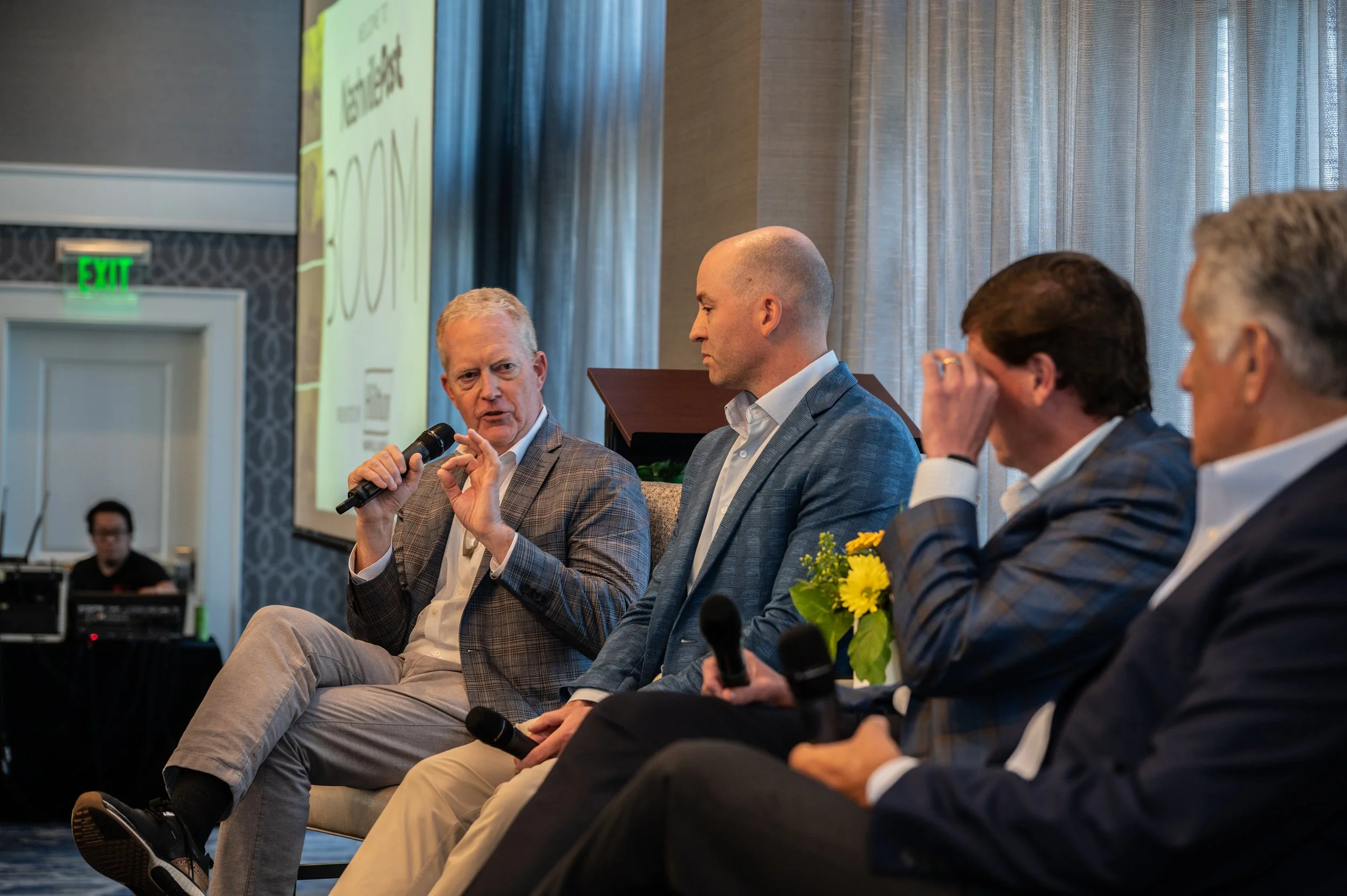 Four men seated on a panel at a conference, with one speaking into a microphone, and a screen in the background displaying text.