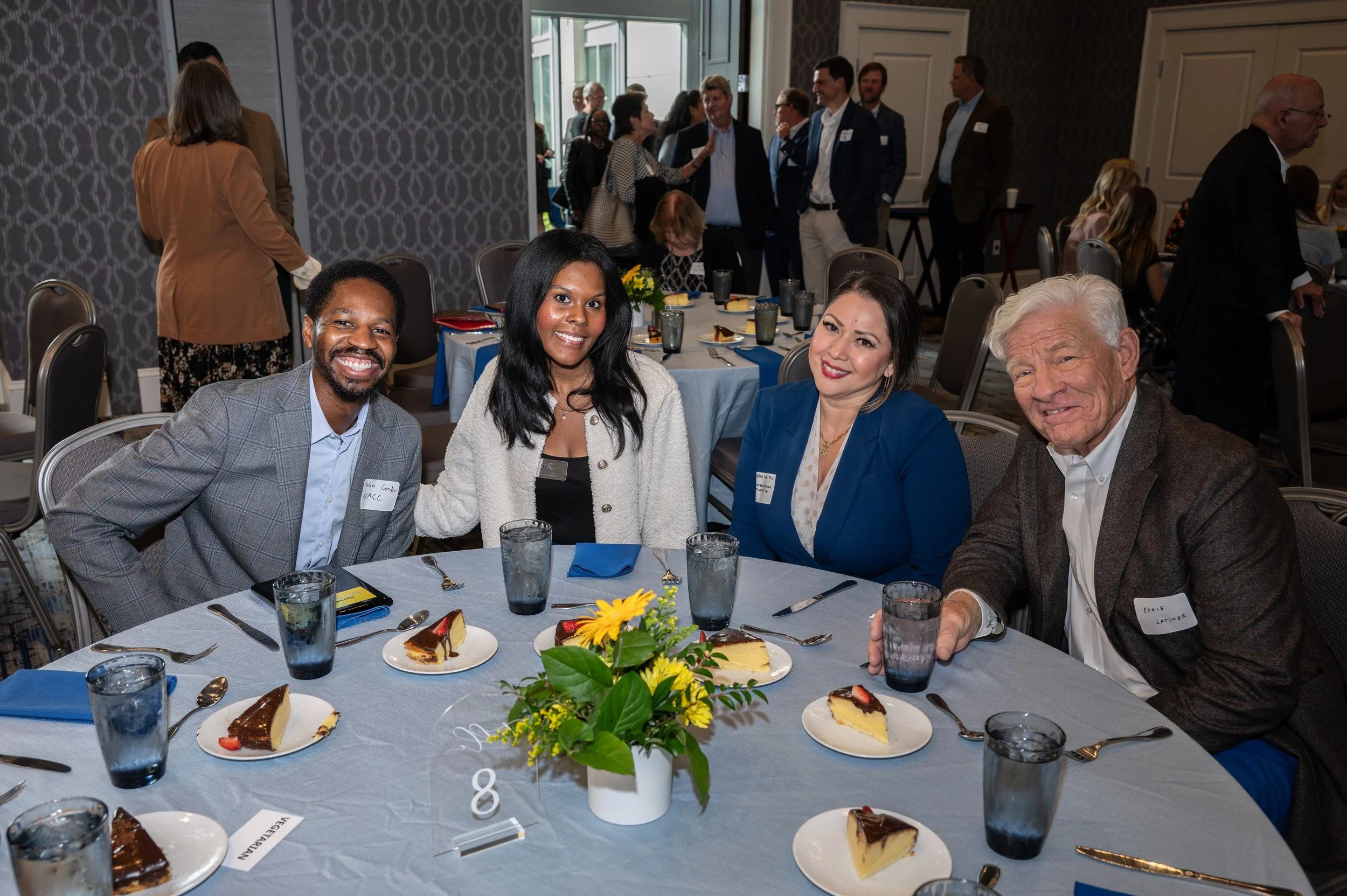 A diverse group of four people sitting at a round table with plates of cheesecake, glasses of water, and a floral centerpiece, smiling at the camera during a social event or conference.