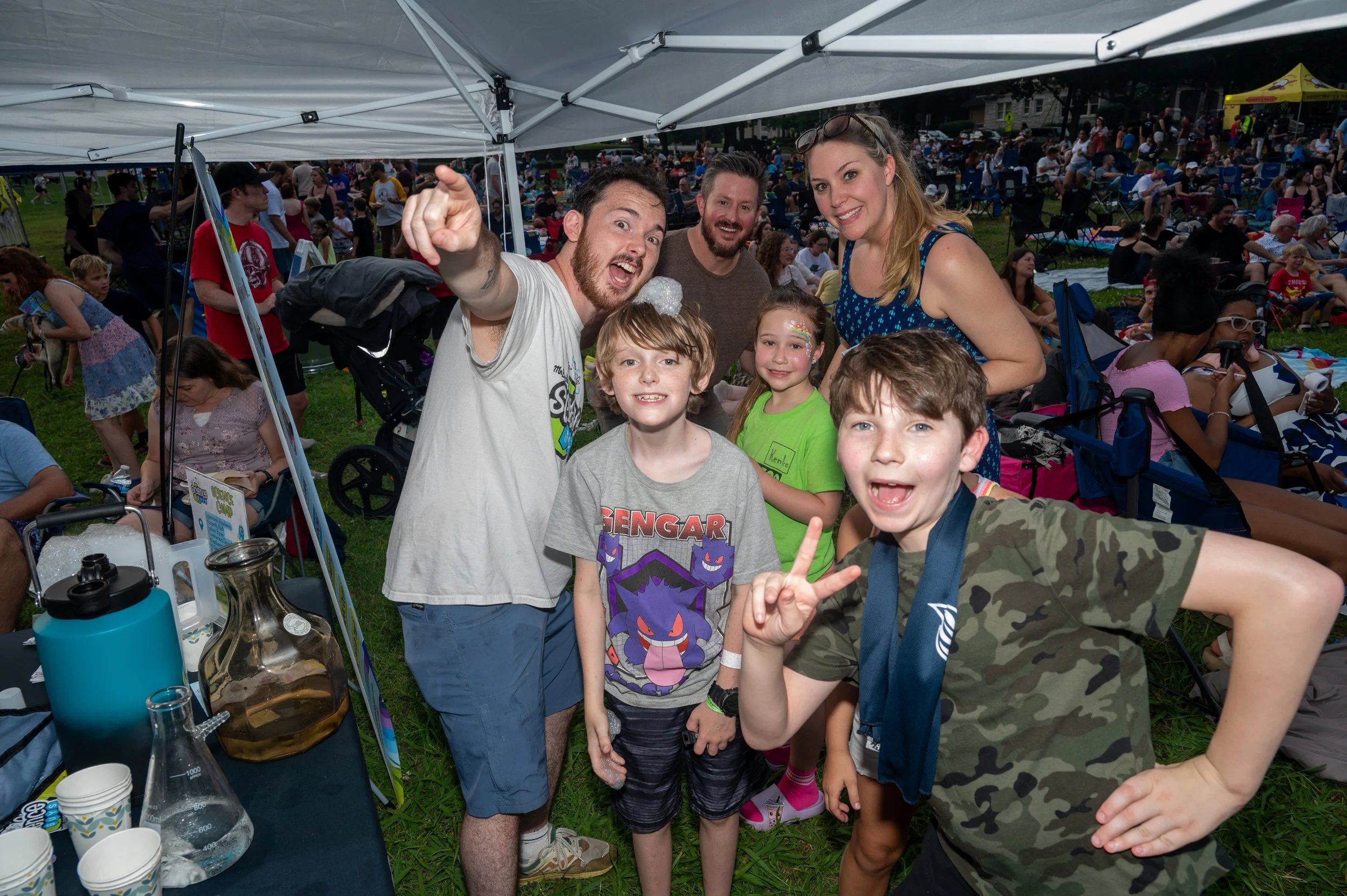 A group of people, including children and adults, gathered under a white canopy at an outdoor event with many people in the background, some seated on the grass and others walking around.