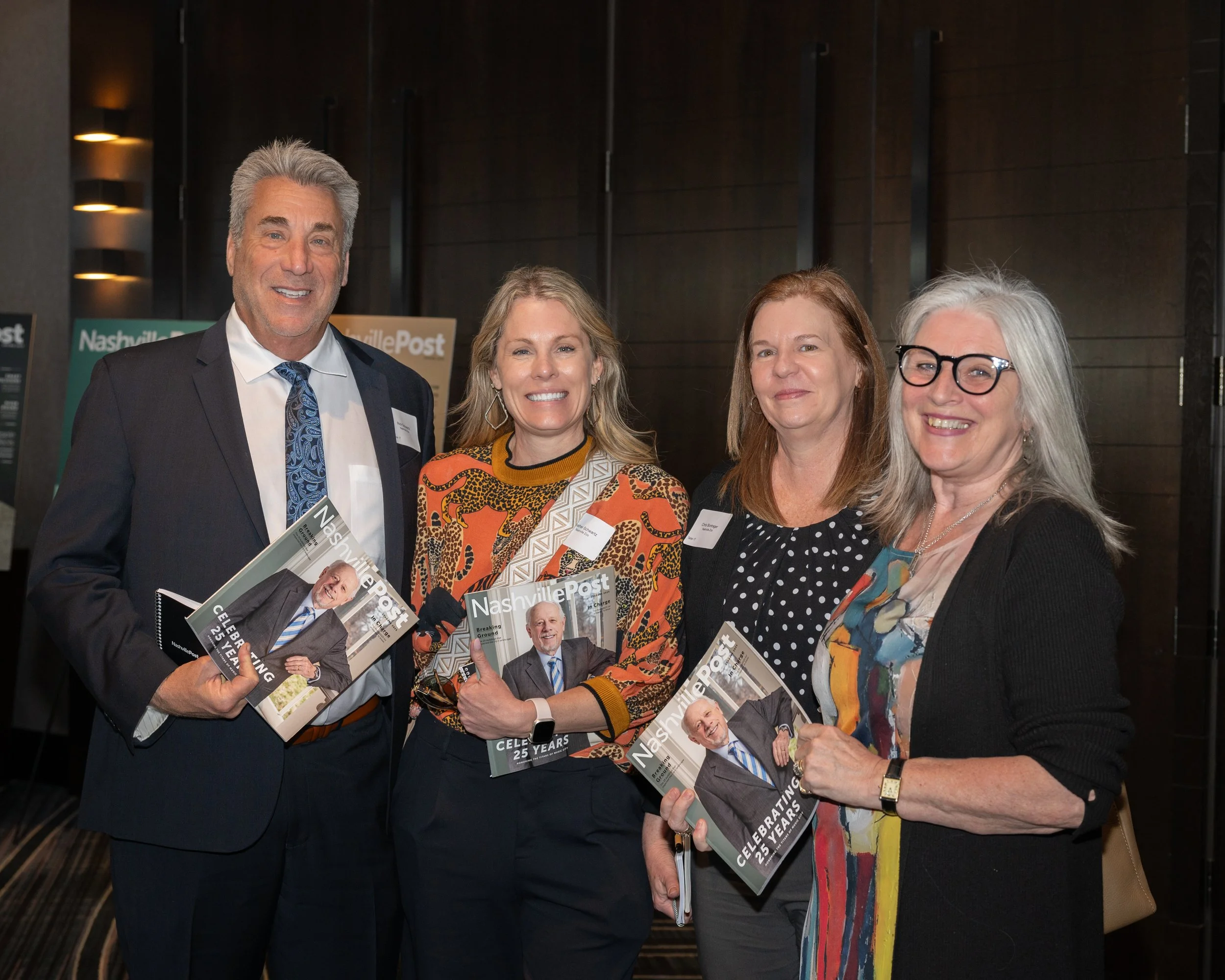Four people standing together at a celebration event, holding Nashville Post magazines, smiling for the camera, against a dark wall background.