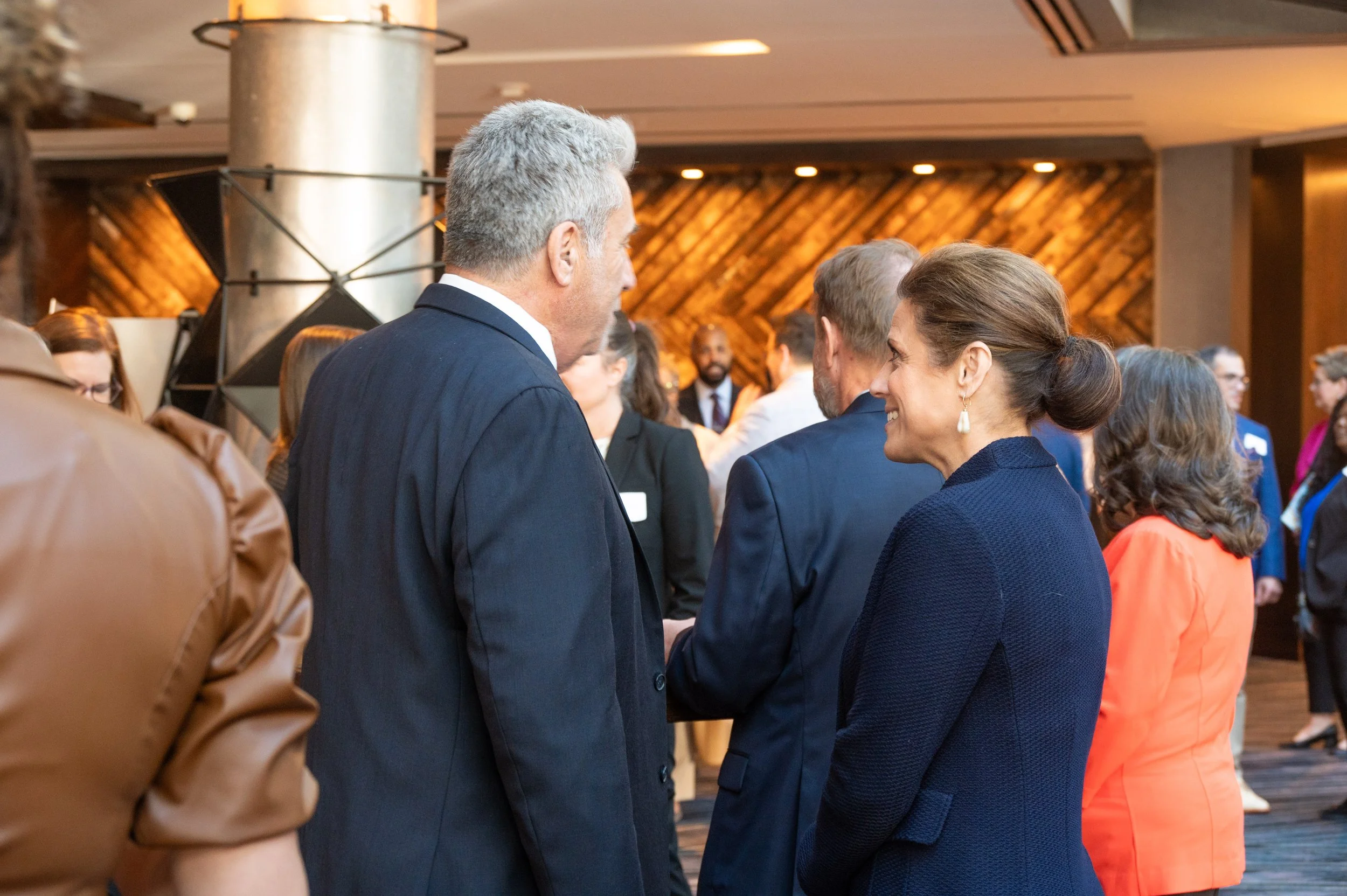 Two professionally dressed individuals, a man and a woman, engaged in conversation at a formal networking event, with other attendees mingling in the background.
