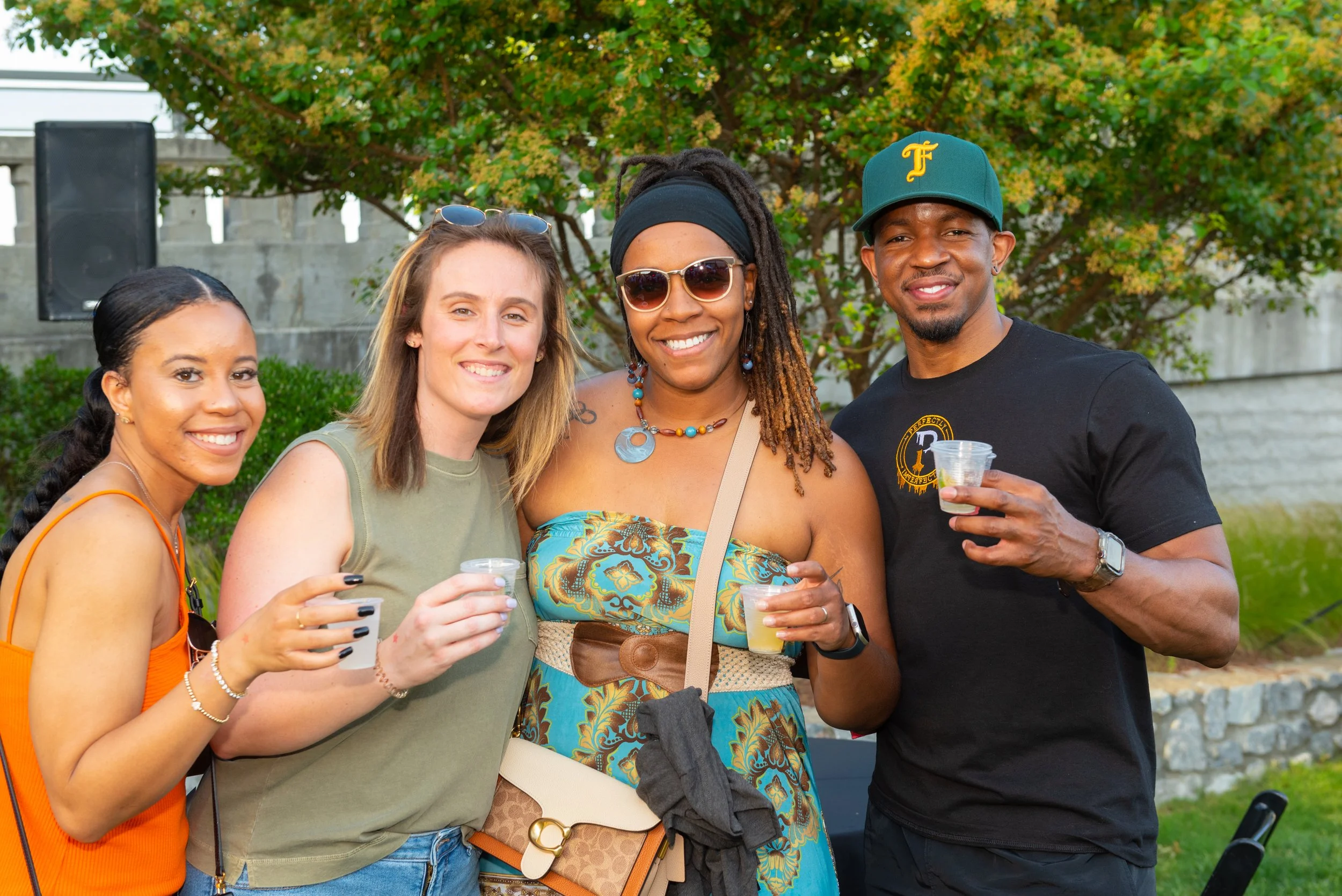 Four friends smiling and holding margaritas at an outdoor gathering, standing in front of a tree with green leaves.