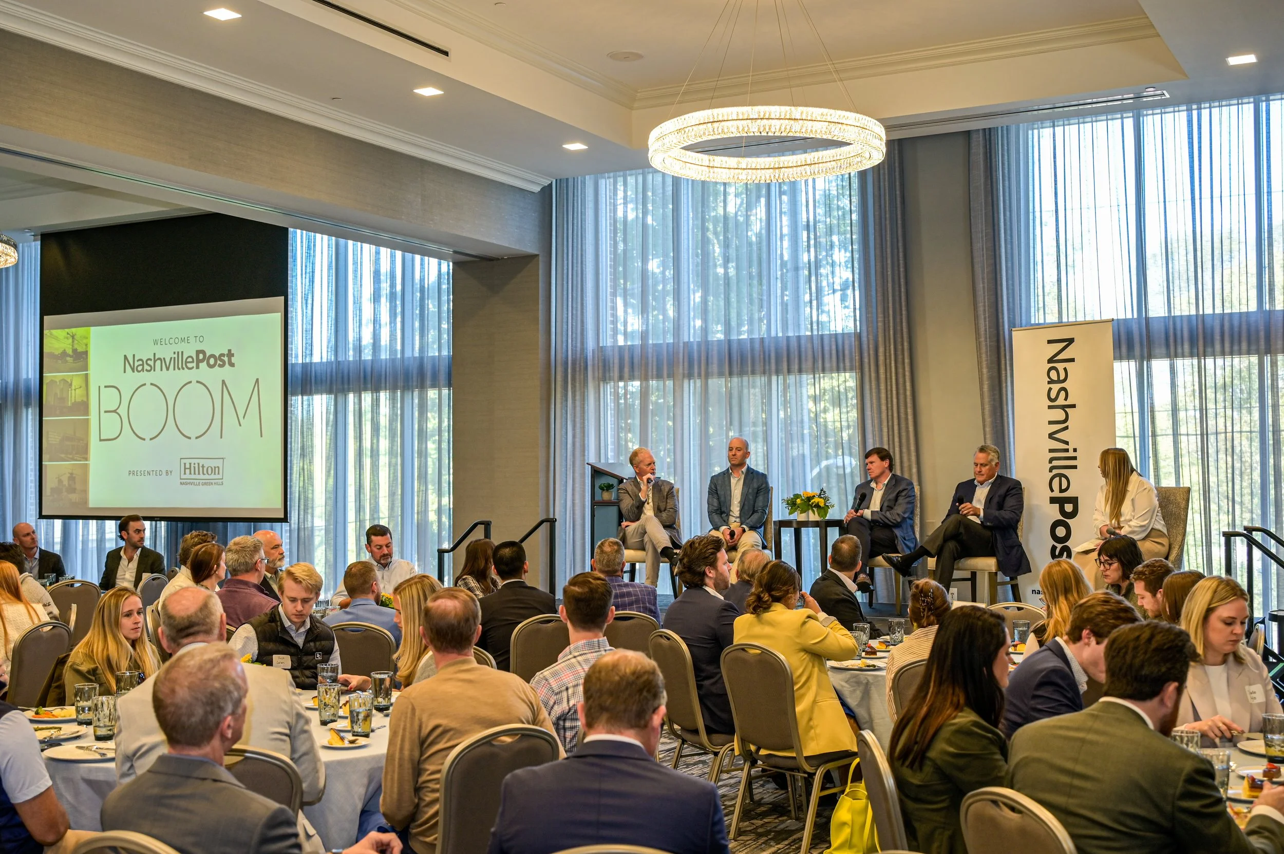 A panel discussion taking place at the Nashville Post BOOM event, with five speakers seated on stage, a large screen displaying the event name, and an audience seated at round tables.