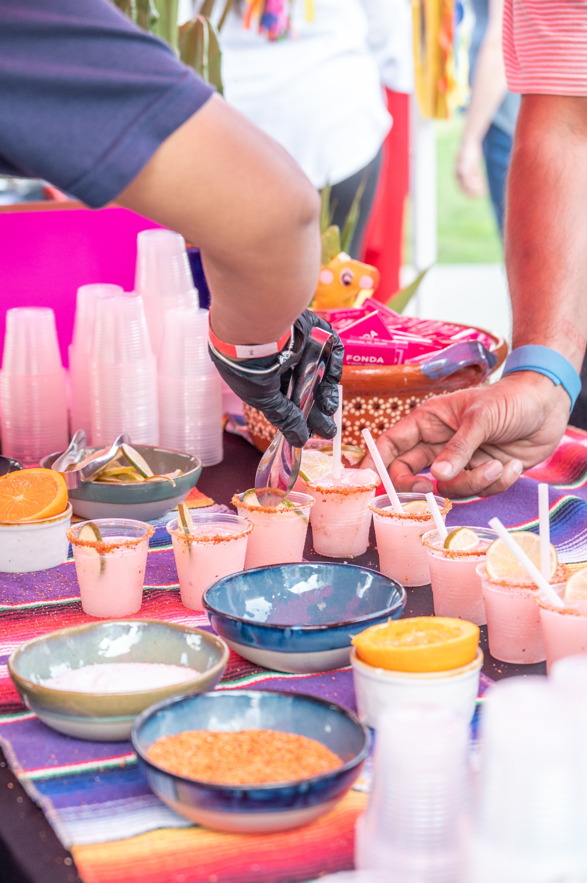 A person is serving pink frozen margaritas with lemon slices to customers at a booth with colorful bowls of ingredients, plastic cups, and a Mexican-themed tablecloth.