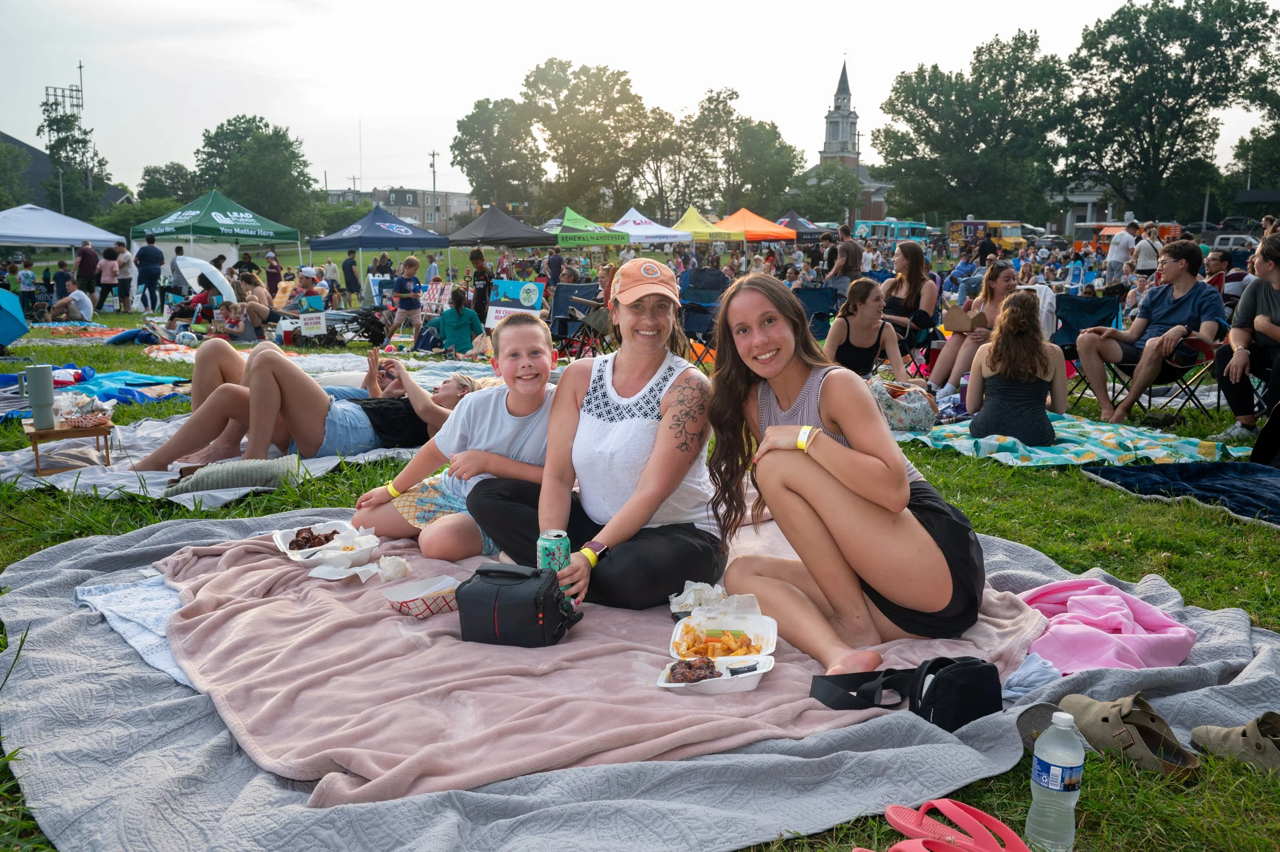 Three people sitting on a blanket at an outdoor event with many tents and people in the background. They are smiling and enjoying food.
