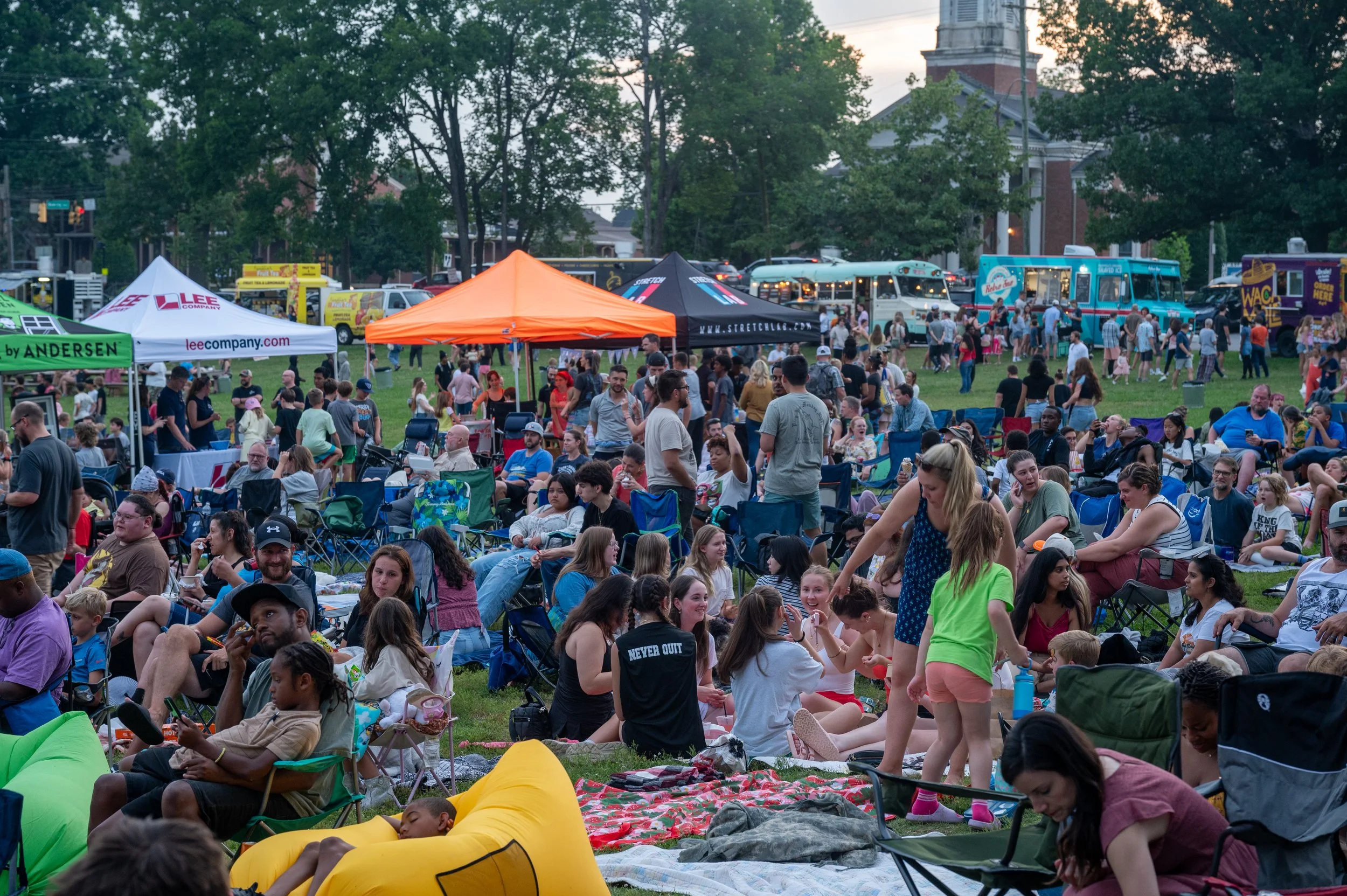 Crowd of people sitting on blankets and folding chairs at Movies in the Park with food trucks and tents in the background.